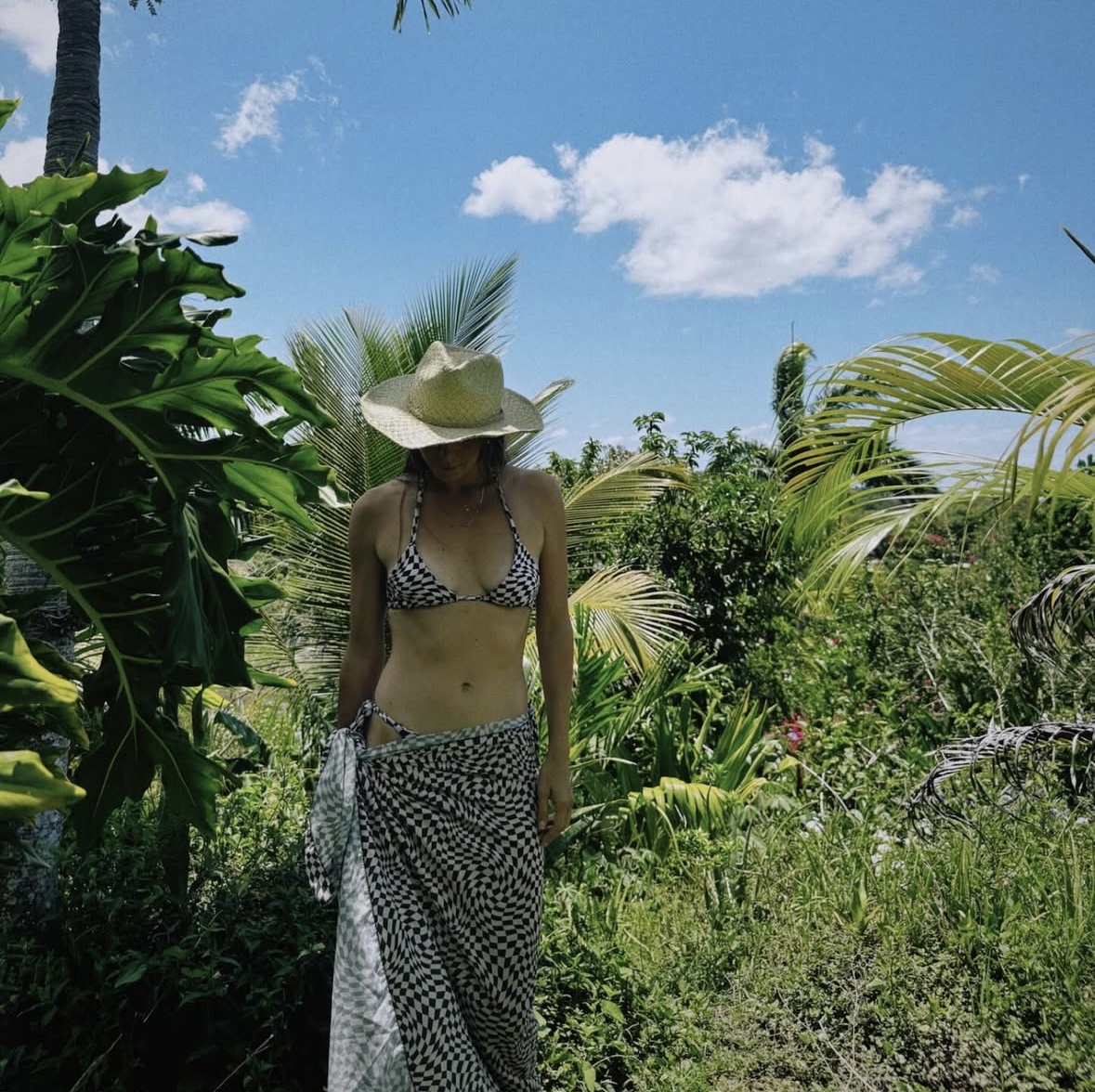 Woman in a bikini and wide-brimmed hat walking through lush tropical greenery under a blue sky with clouds.