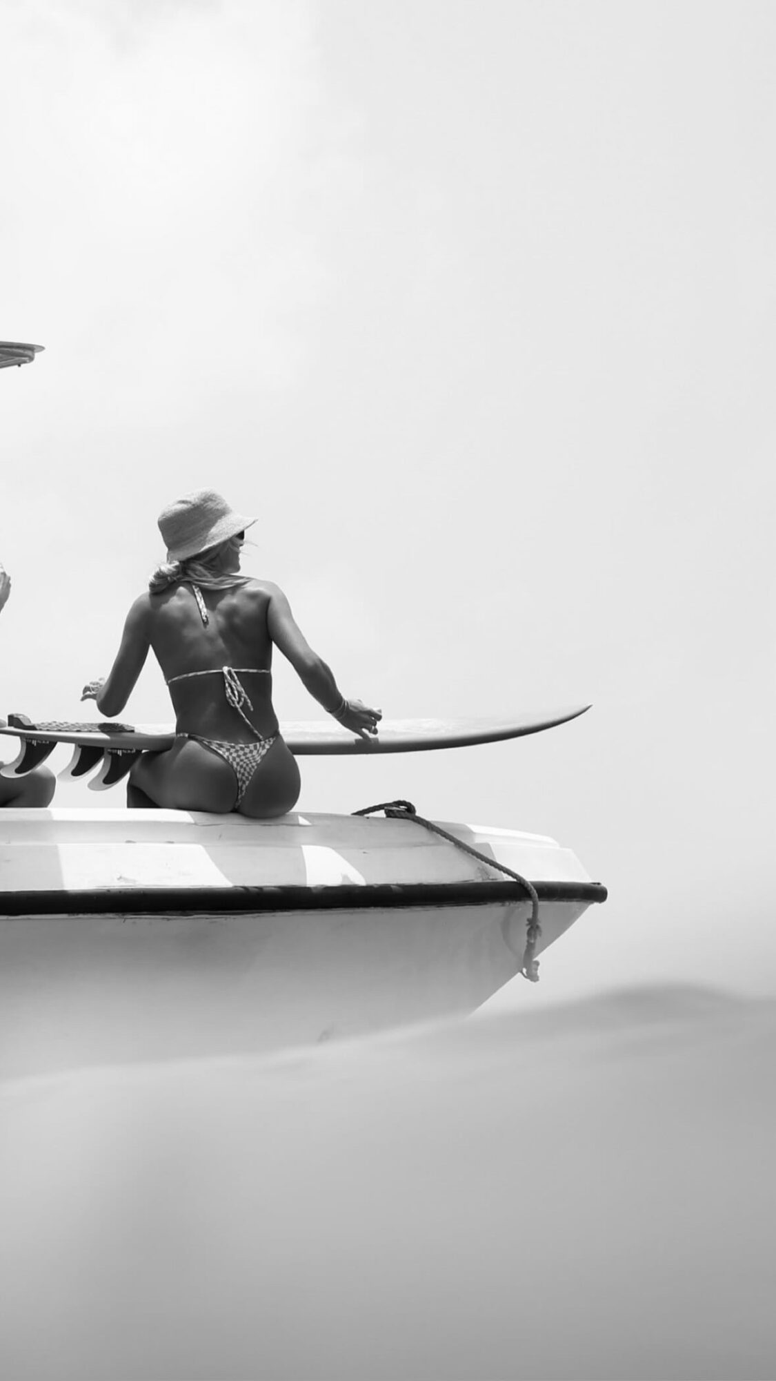 Person sitting on a boat holding a paddle, wearing a hat, with a kayak nearby, in black and white.