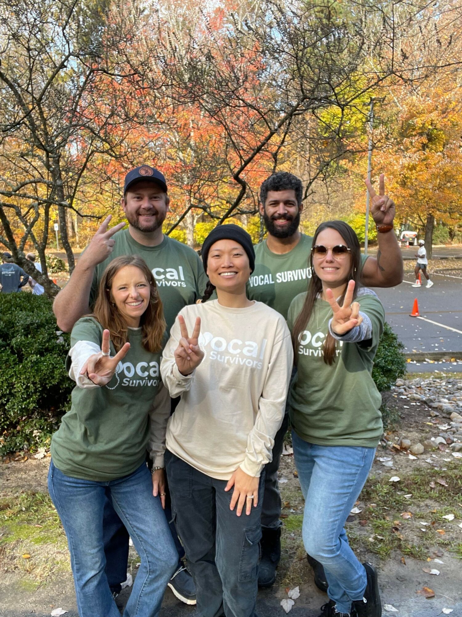 Group of five people outdoors with autumn trees, some making peace signs, smiling, wearing casual clothing.