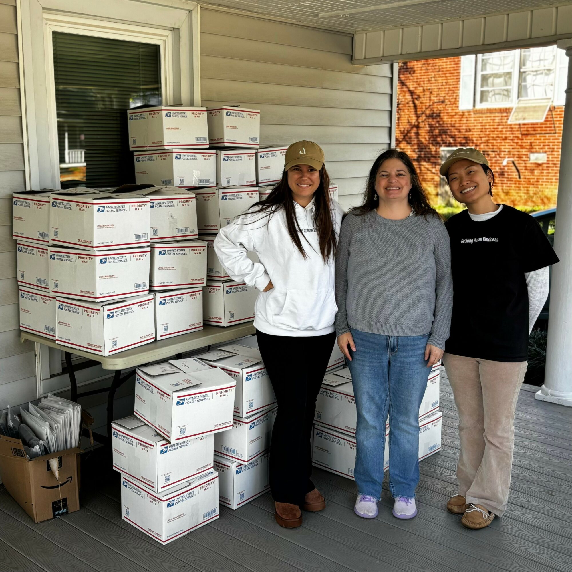 Three people stand on a porch with boxes stacked behind them, smiling at the camera, autumn trees visible in the background.