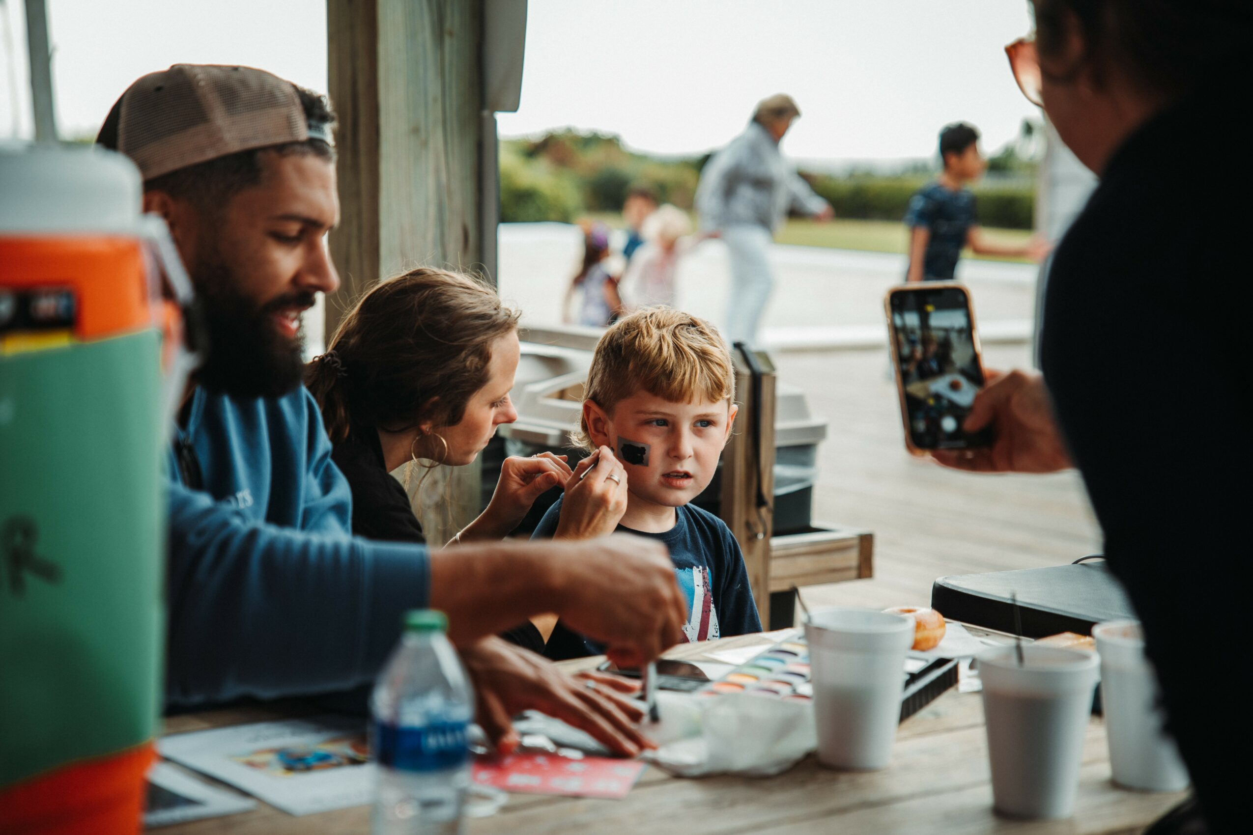 Group of people, including children, sitting outdoors at a table with drinks and snacks, with others walking in the background.