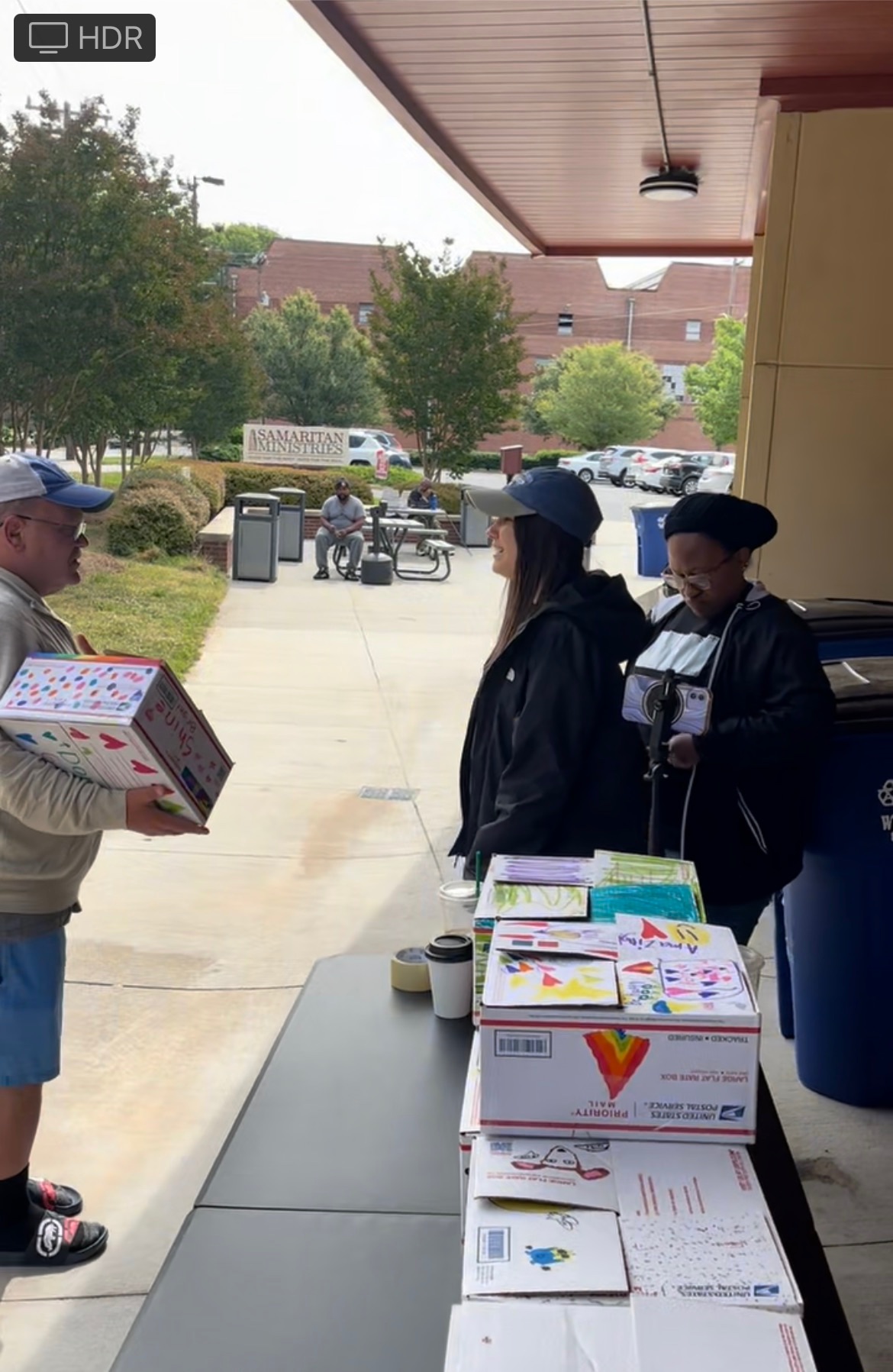 Person in gray shirt and blue shorts holding a pizza box, talking to two people in dark clothing outside a building, with trees and parked cars in background.