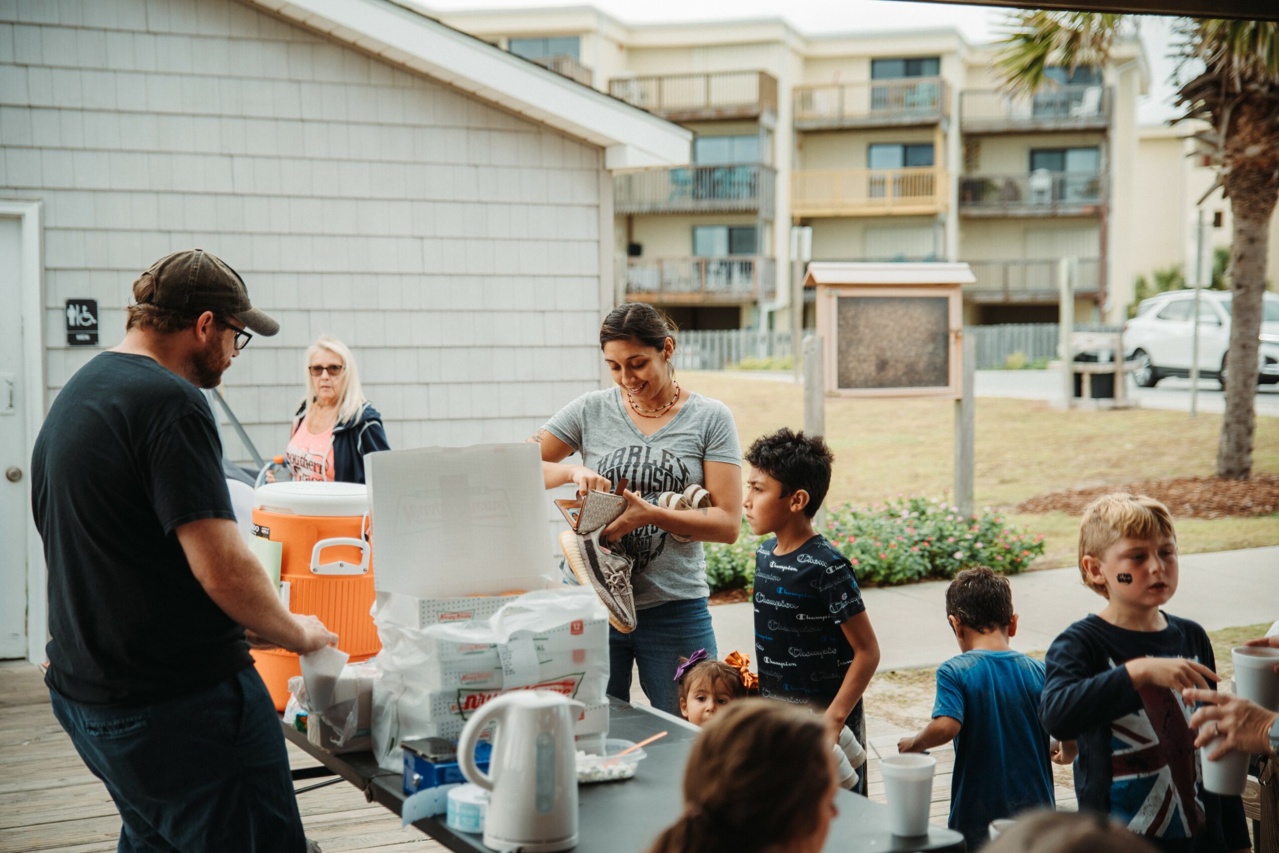 Group of children and adults outdoors near a building, with a woman handing out items and a man in black shirt nearby.