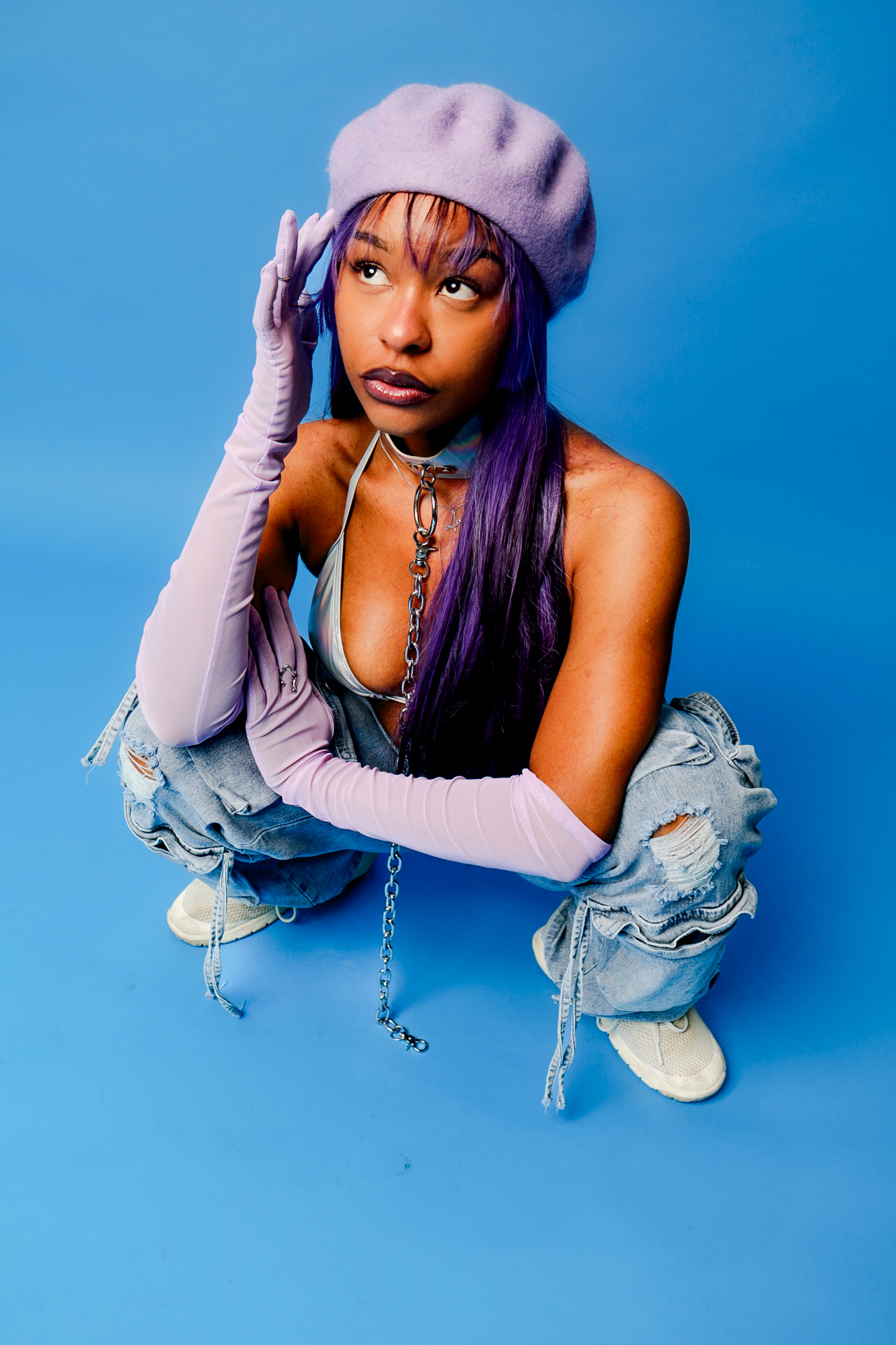 Young woman with purple hair wearing a lavender beret and long gloves, squatting against a blue background.