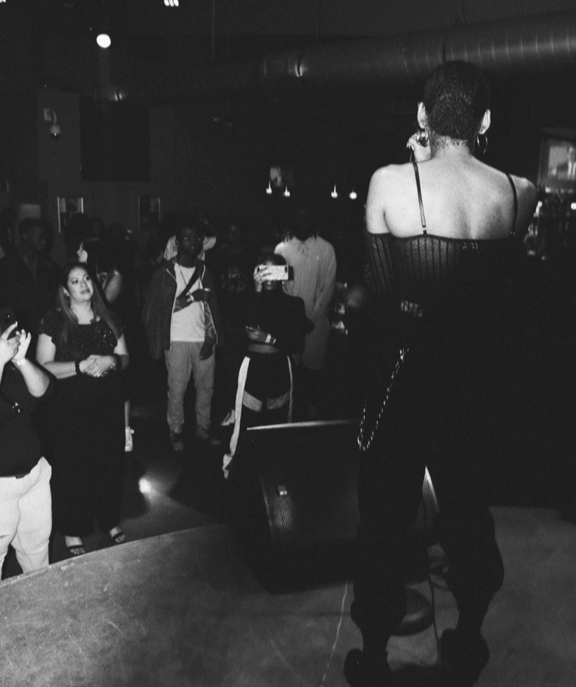 Performer on stage with back to camera, audience watching in a dark venue, black and white photo.