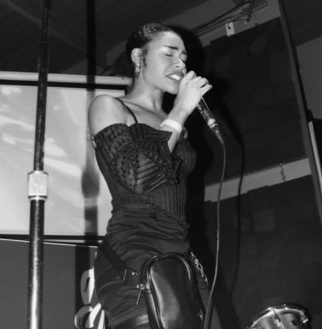 Woman singing into a microphone with eyes closed, wearing a dark off-shoulder top, in black and white.