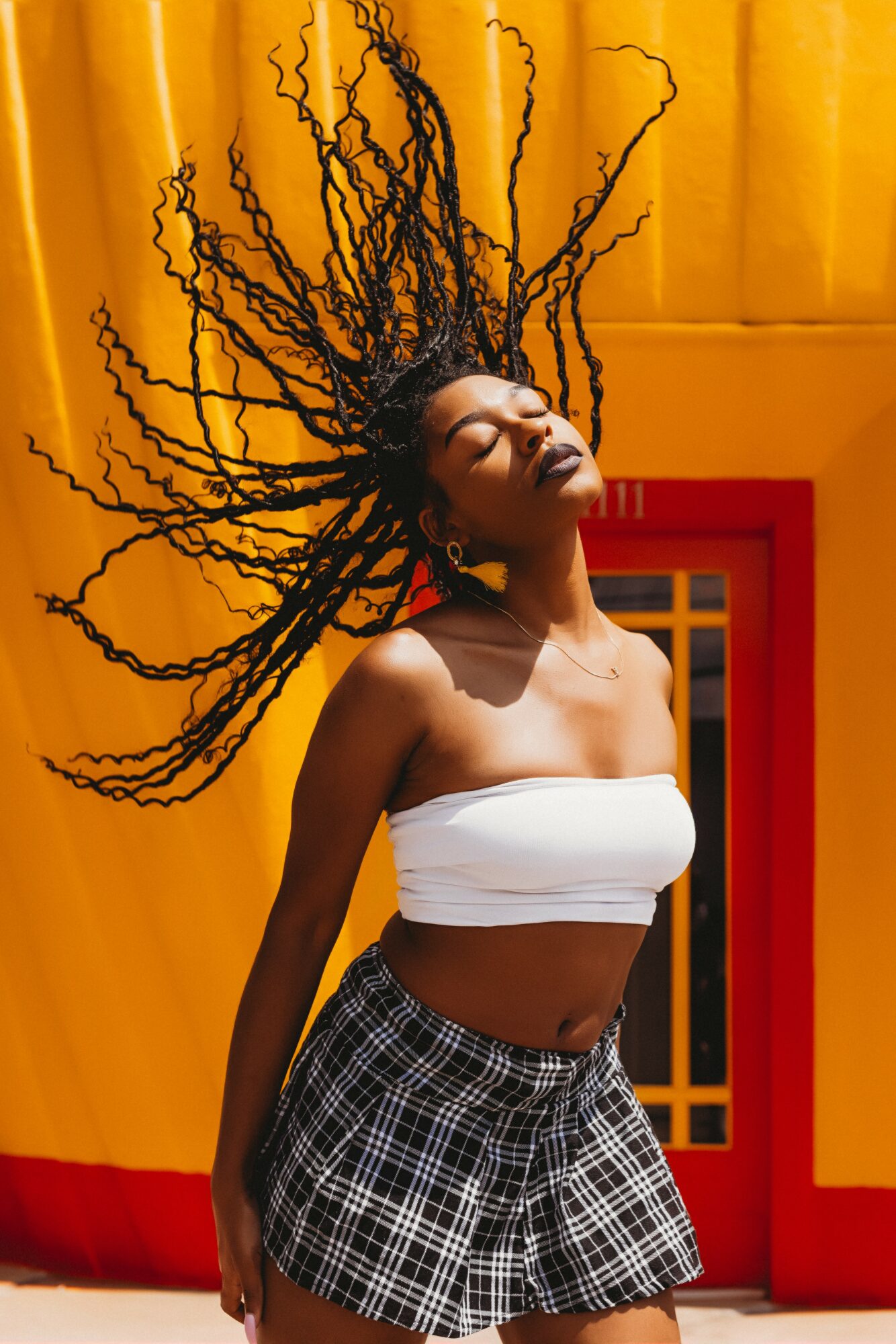 Woman with long, curly hair outdoors, eyes closed, wearing a white tube top and plaid shorts, yellow background.