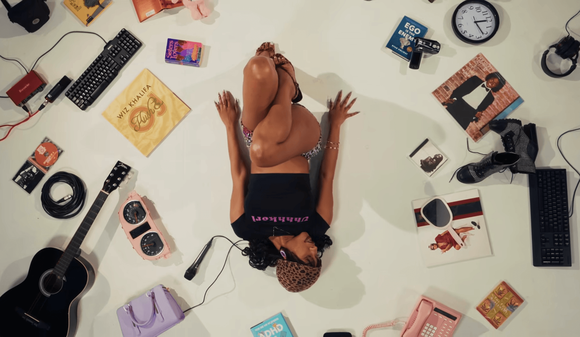 Person lying on floor surrounded by various objects including a guitar, books, and electronic devices.
