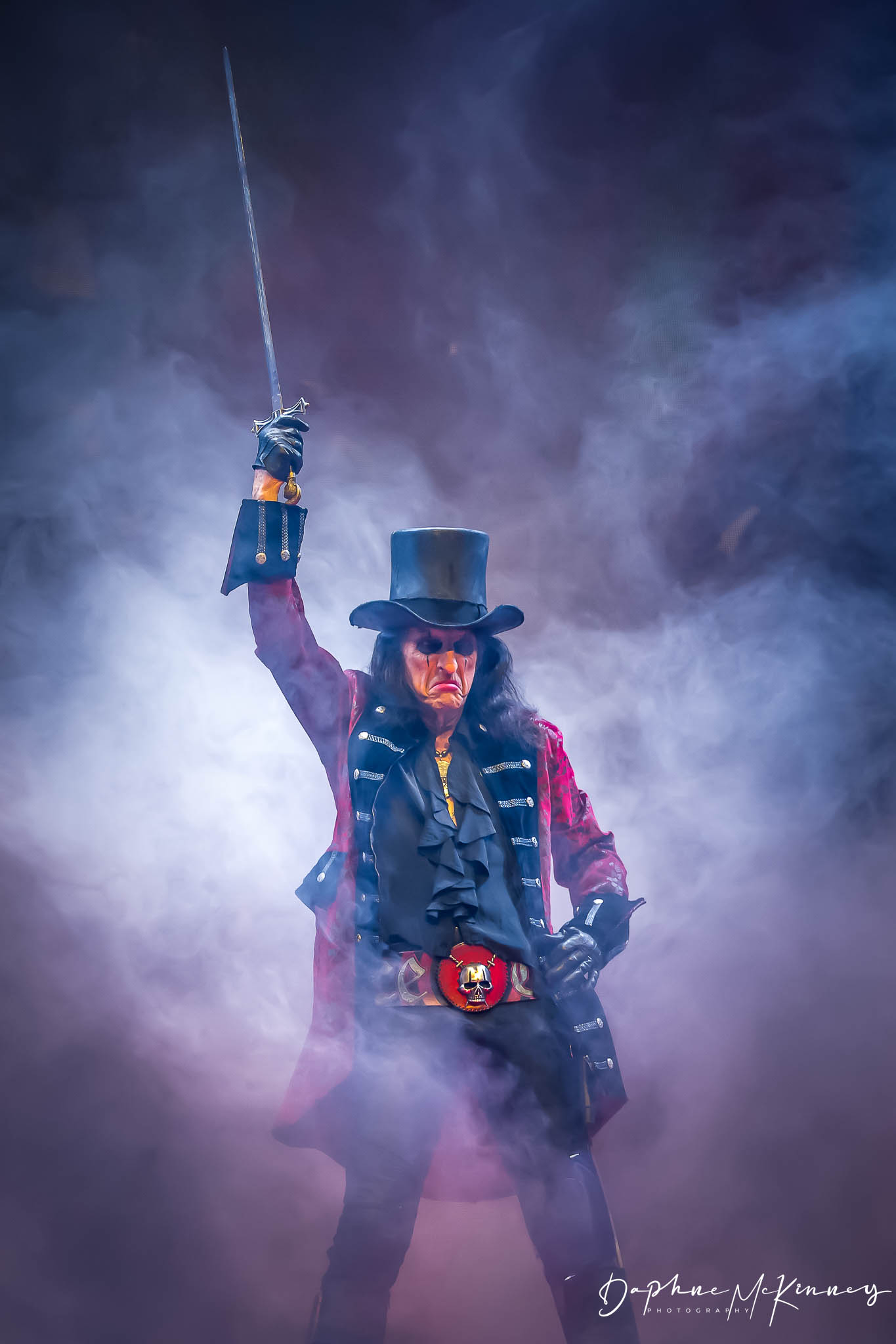 Person dressed as a magician or performer in a top hat, sunglasses, and colorful clothing, holding a wand in a smoky environment.