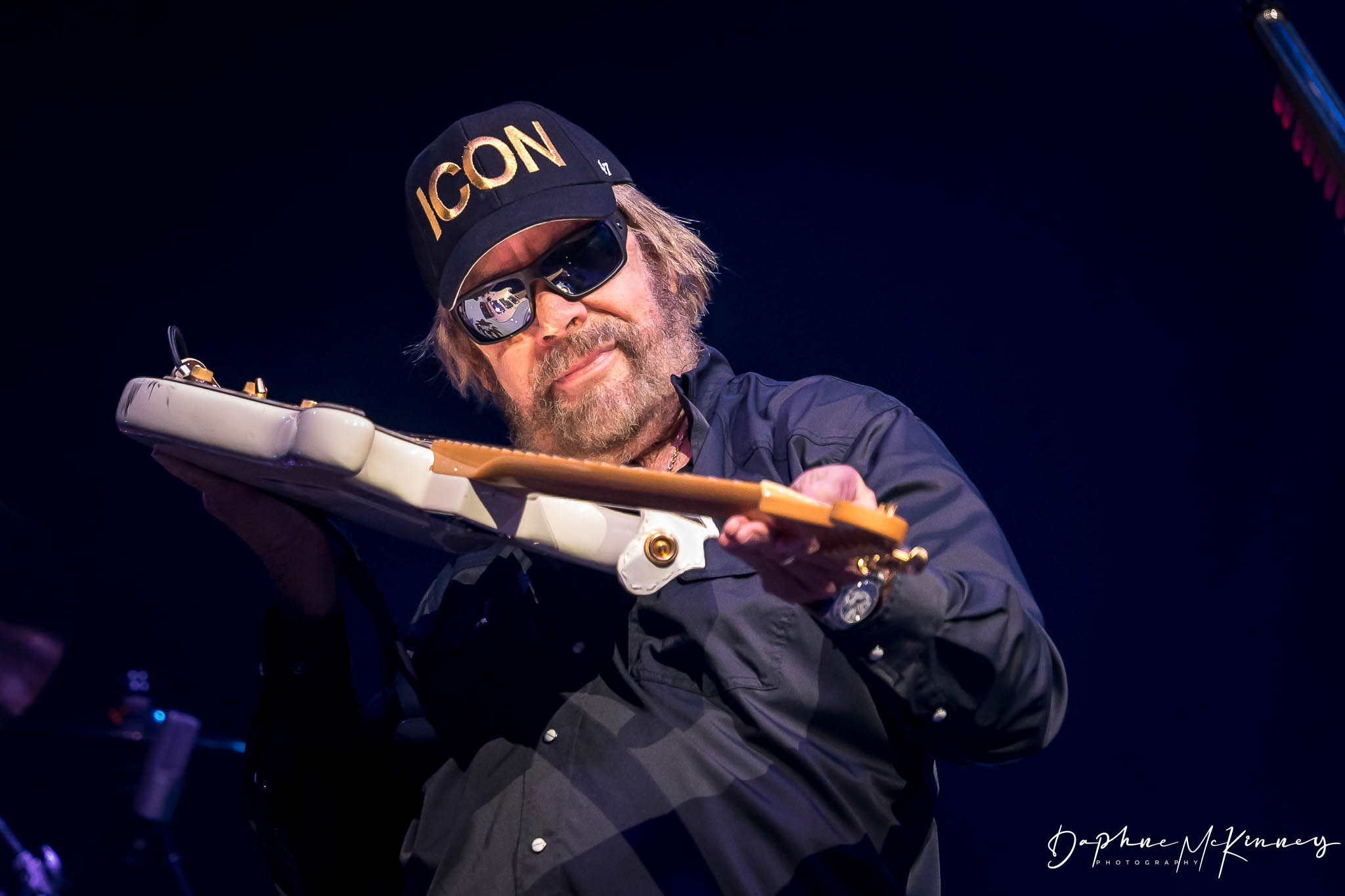 Person with sunglasses and a hat playing a guitar on stage, dark background, stage lighting, casual clothing