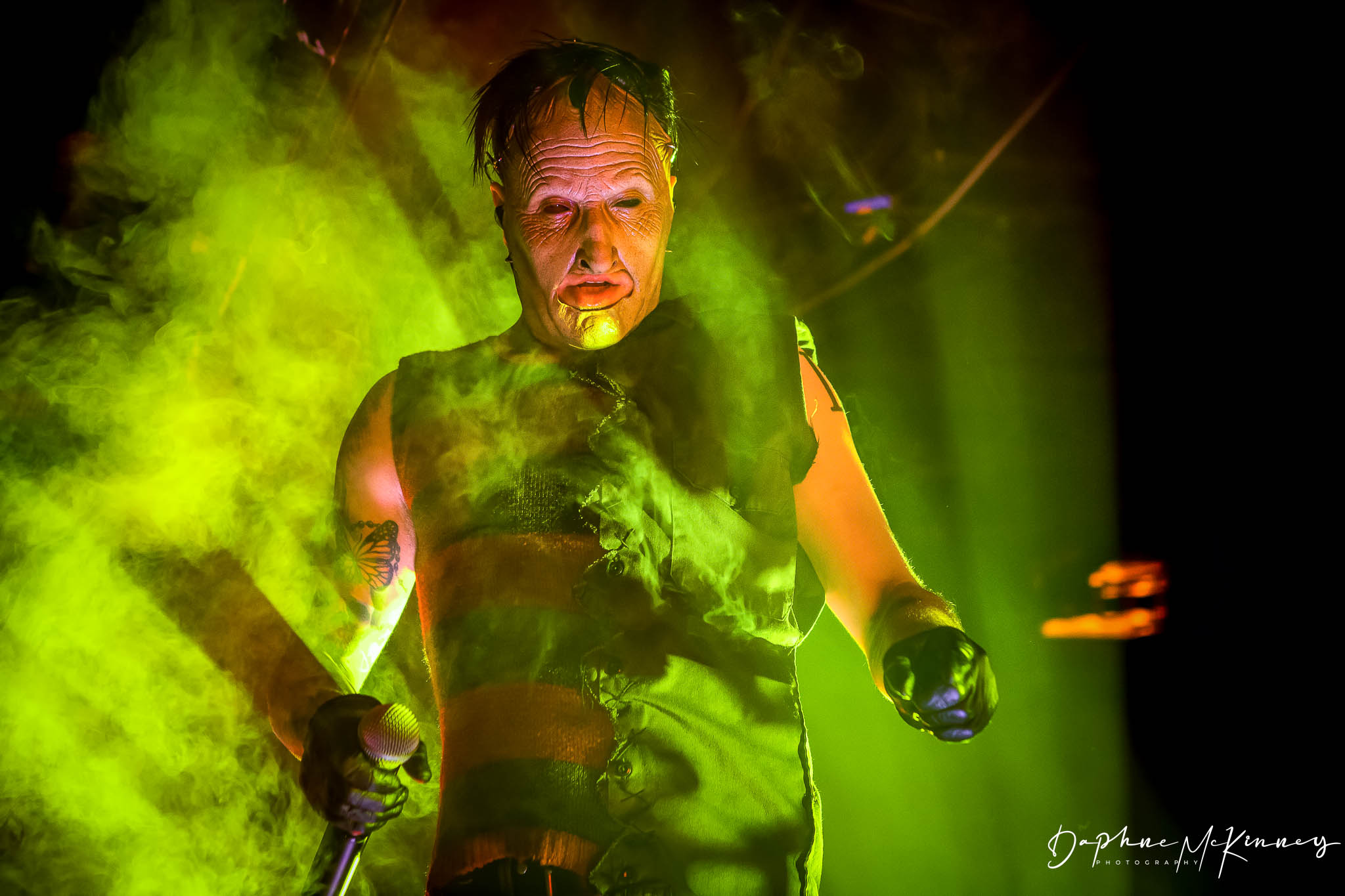 Person with face paint and gloves, surrounded by green smoke and dramatic lighting, holding objects in both hands.