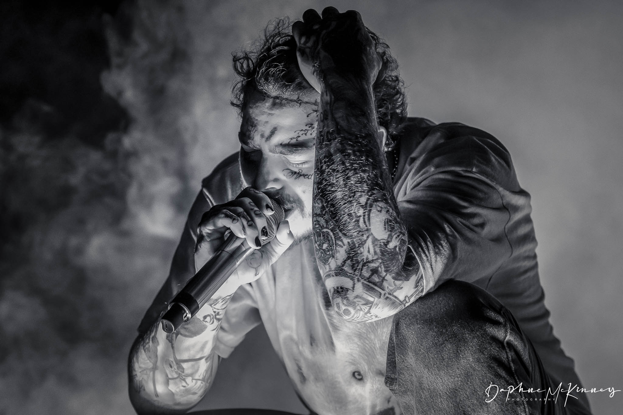 Person singing into a microphone with closed eyes, hand on head, in black and white, with smoke in background.