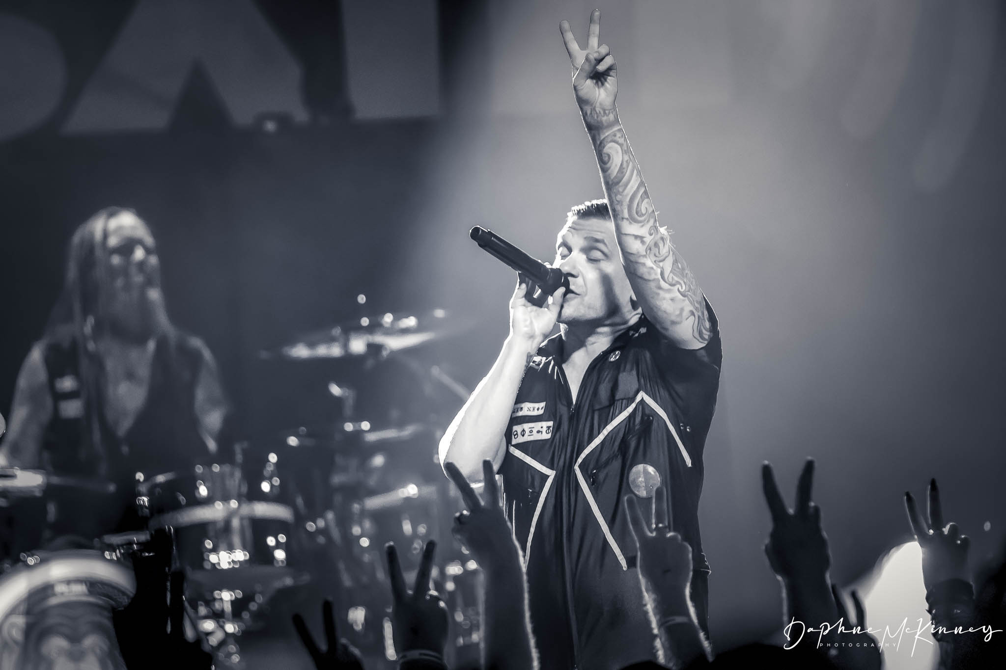 Performer singing into microphone with raised hand, tattoos visible, audience making peace signs, black and white photo.