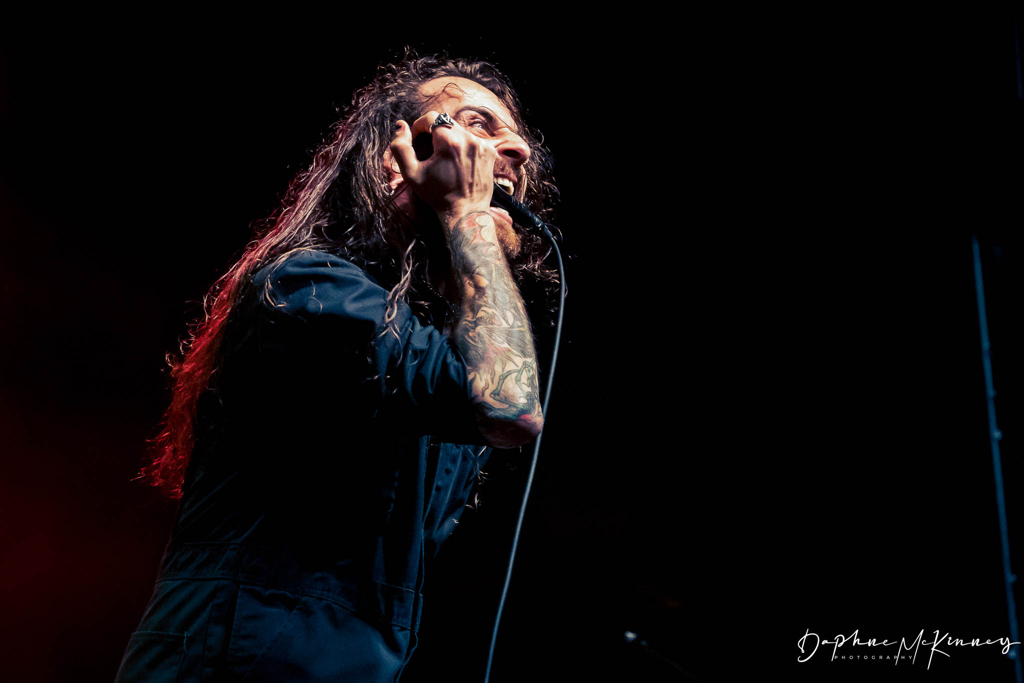 Person singing into a microphone with long hair and tattoos, dark background, stage lighting.