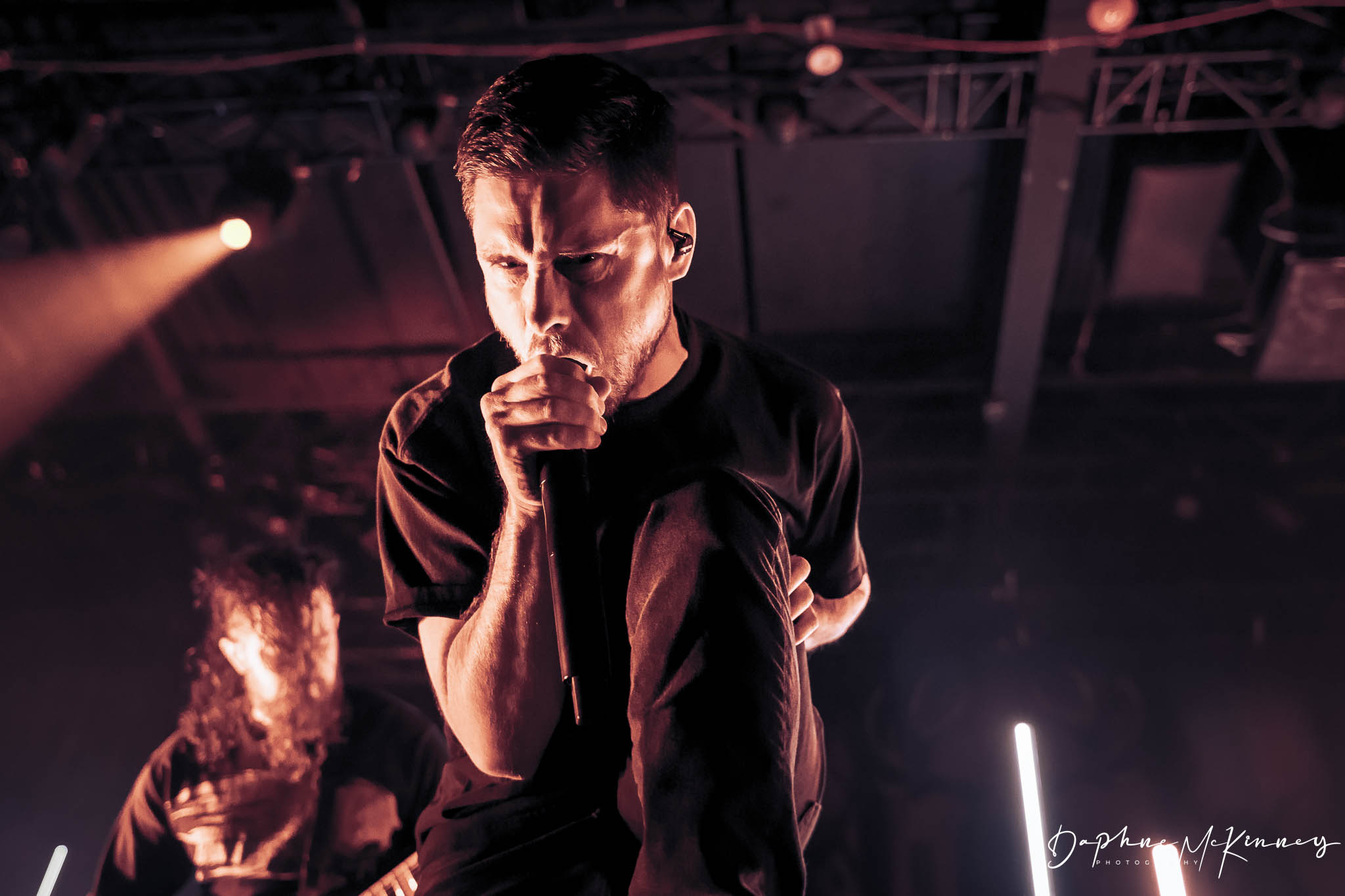 Male singer performing on stage, holding microphone, with a person in the background, stage lighting visible.