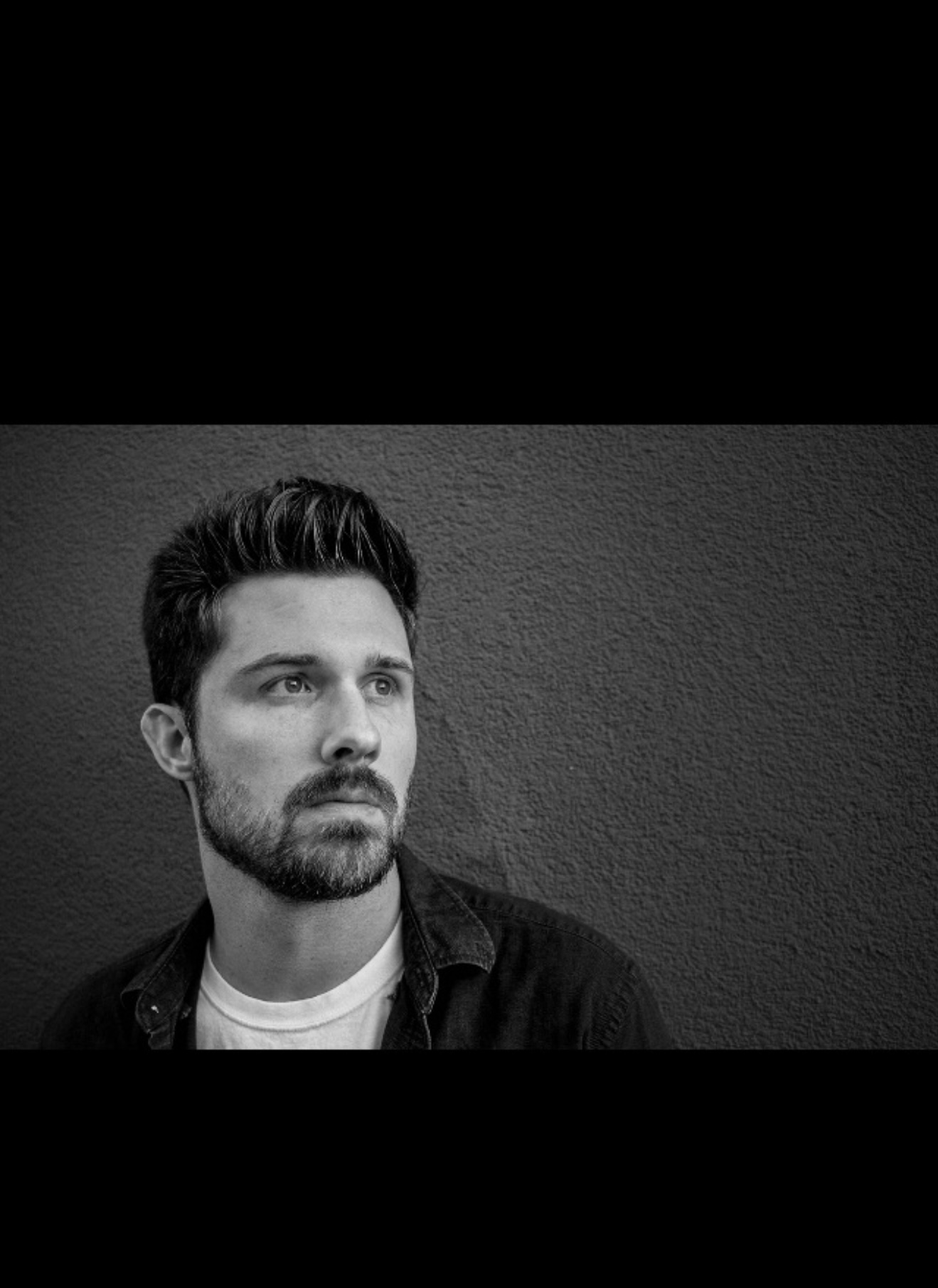 Black and white portrait of a man with short hair and a beard, looking to the side against a textured wall.