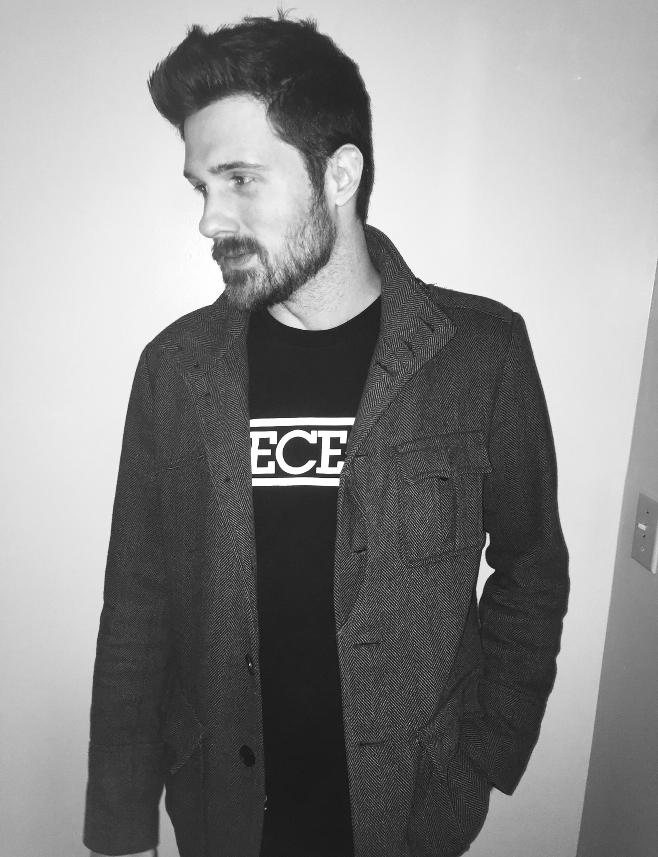 Man with dark hair and beard wearing a dark jacket and T-shirt, looking to the side, standing indoors.