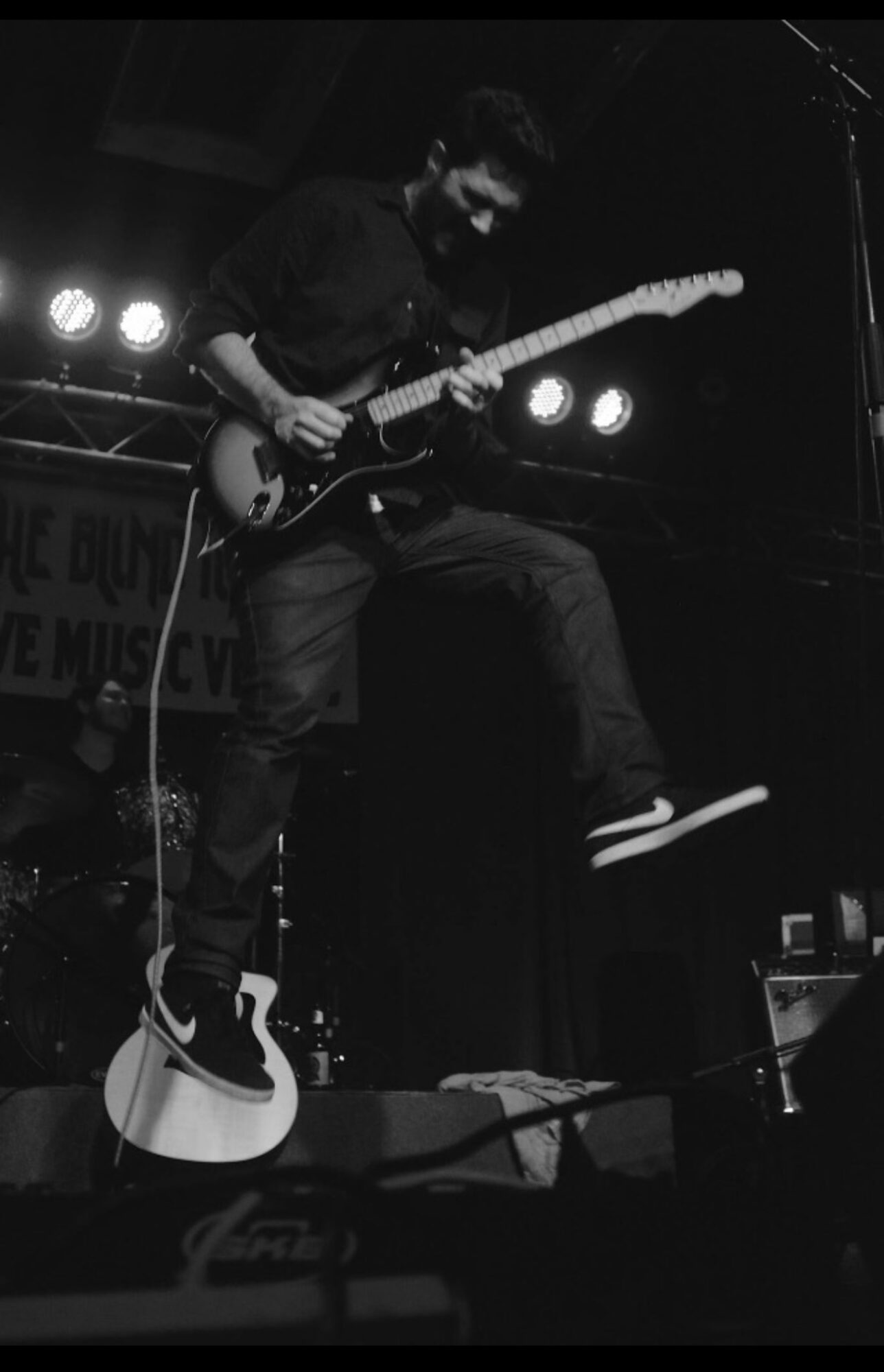 Person playing electric guitar on stage, mid-air jump, stage lights in background
