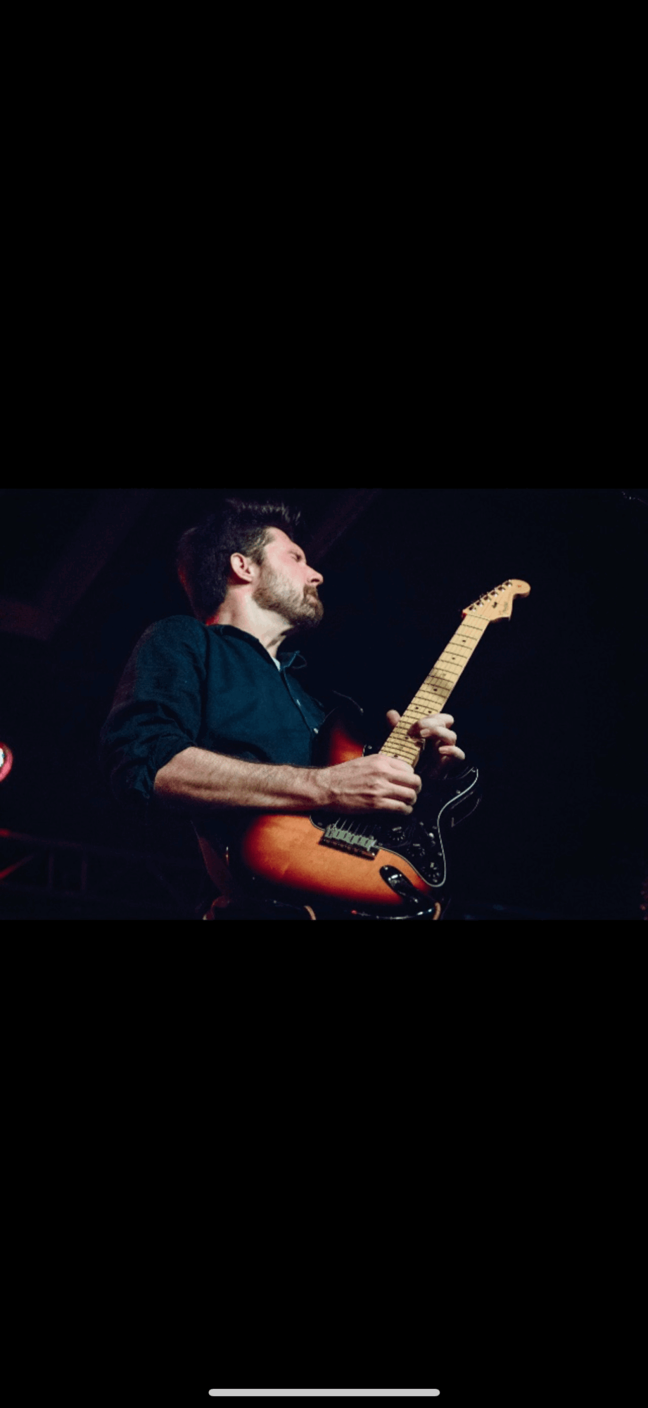 Person playing electric guitar on stage with dark background, wearing dark clothing, focused on performance.