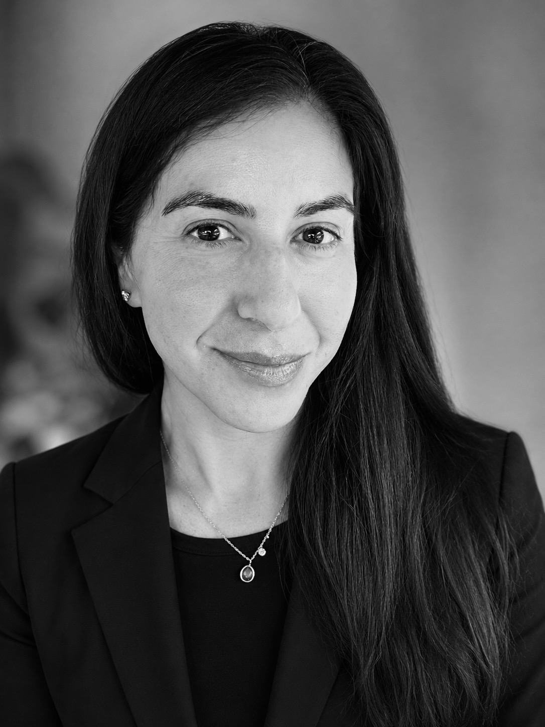 Black and white portrait of a woman with long dark hair, smiling, wearing a blazer and a necklace.