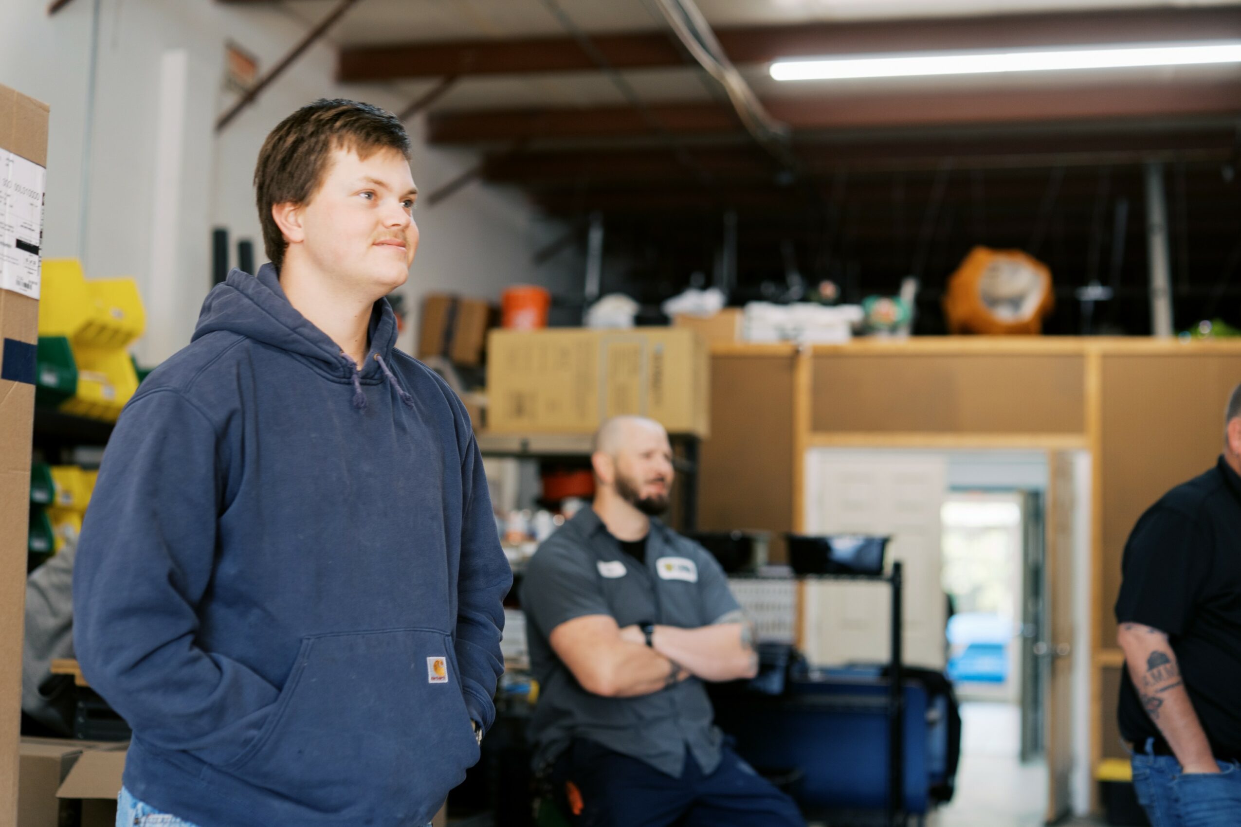Young man in a blue hoodie standing with arms in pockets, smiling, in a warehouse setting with shelves and boxes.
