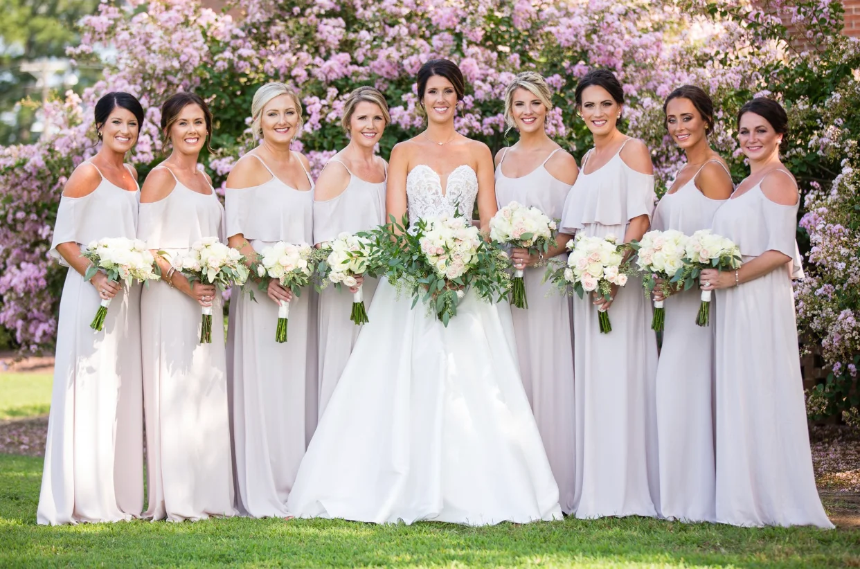 Group of nine women in white dresses holding bouquets, standing outdoors in front of pink flowering bushes.