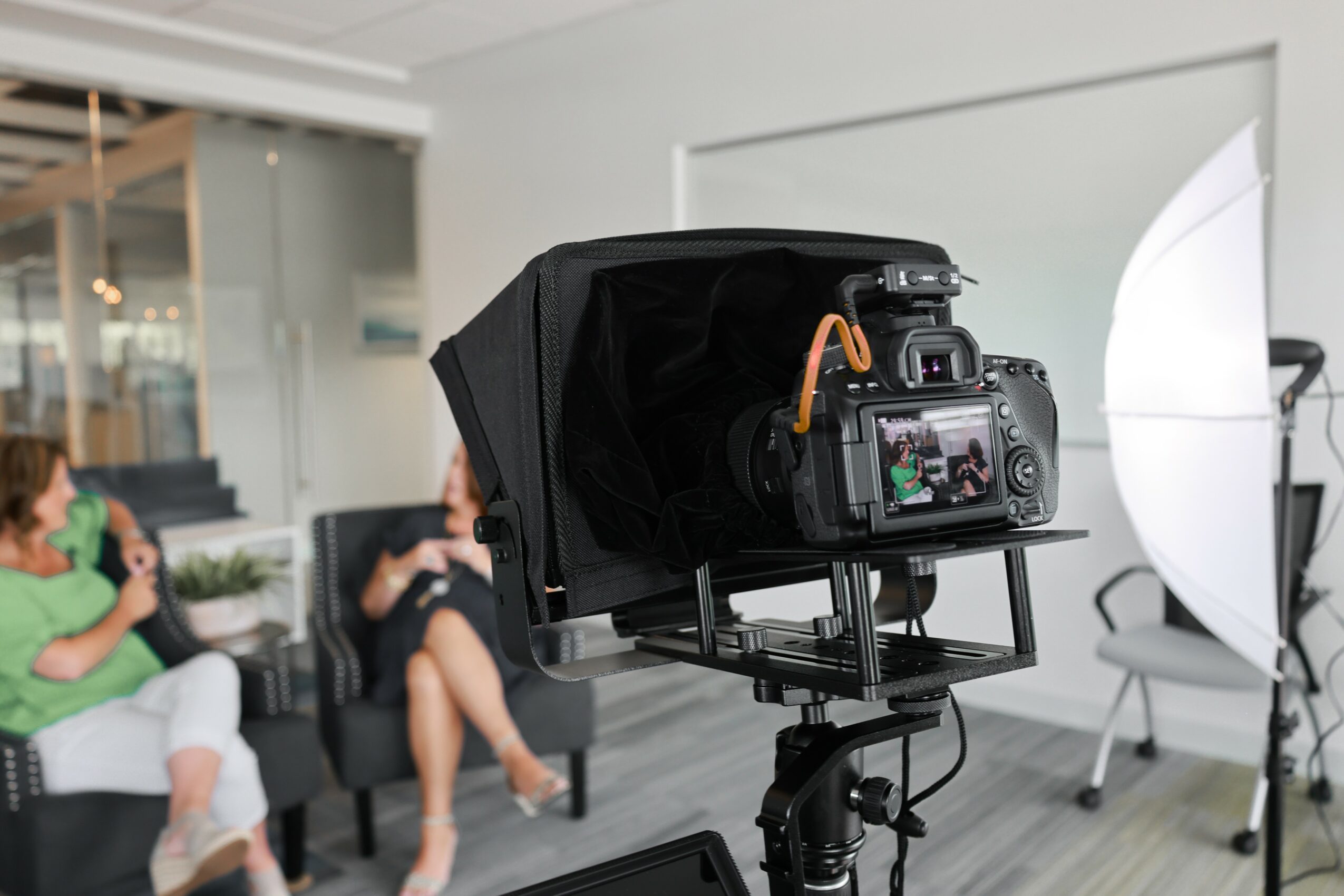 Photography studio setup with camera, softbox, and two people sitting on chairs in background.