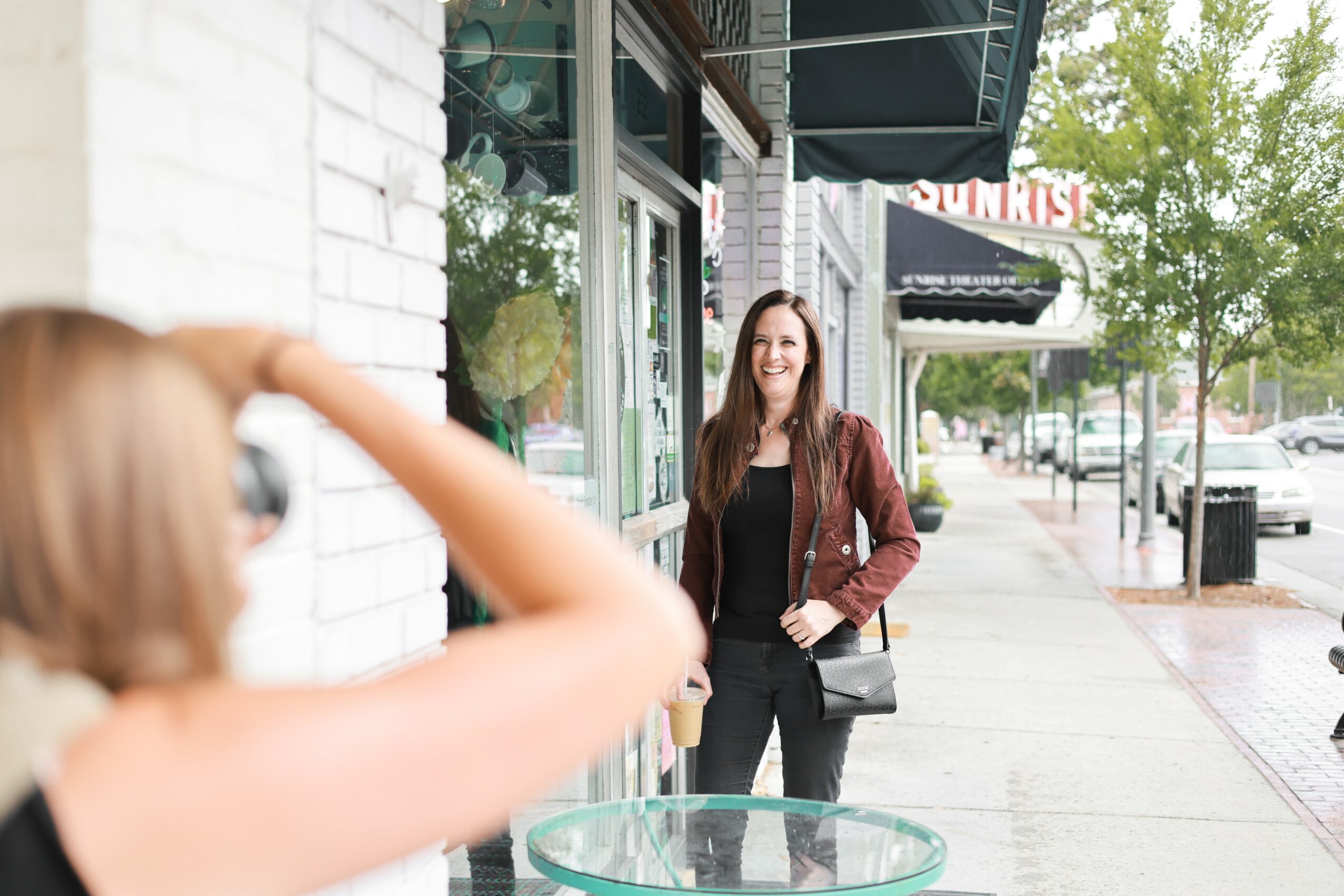 Woman with long brown hair standing on sidewalk outside store, holding a bag, smiling, with trees and parked cars in background.
