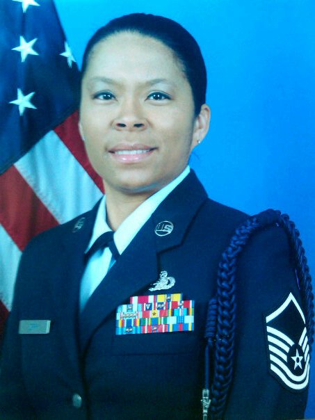 Female military officer in uniform with ribbons, standing in front of an American flag, smiling.
