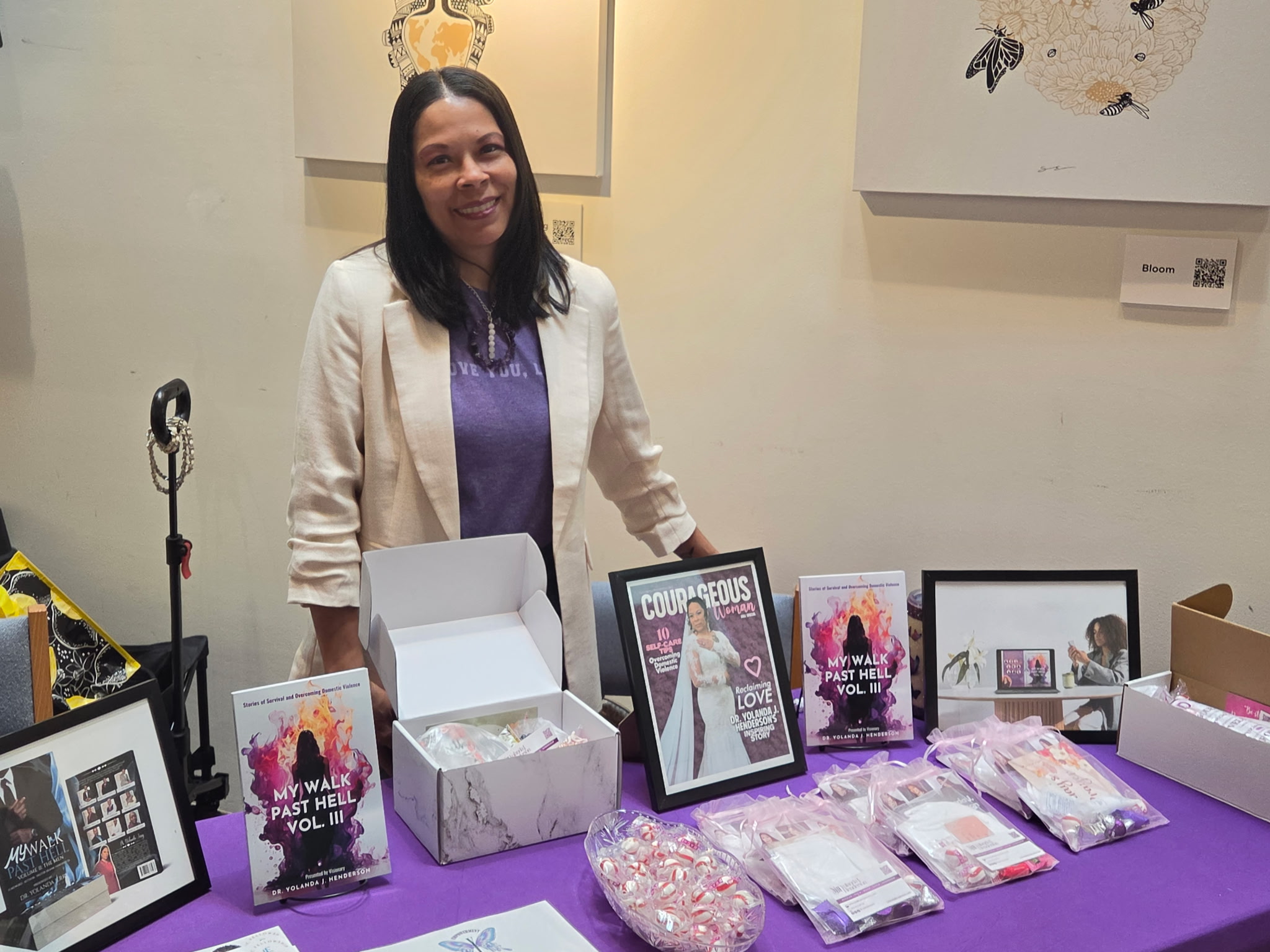 Woman standing behind a table with framed pictures and packaged items, smiling, in an art gallery setting.