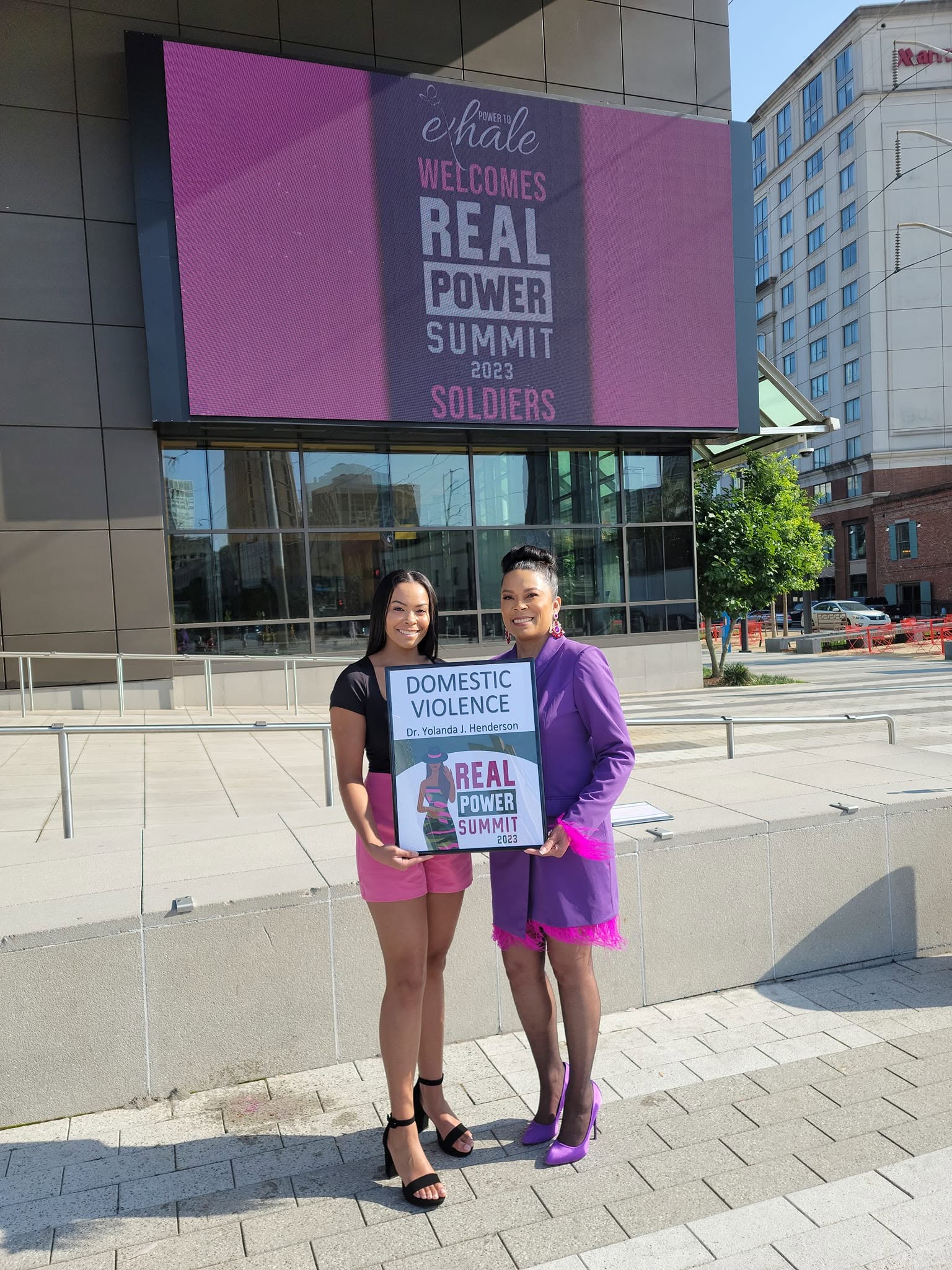 Two women stand outside a building holding a sign about domestic violence awareness, with a large digital screen behind them.