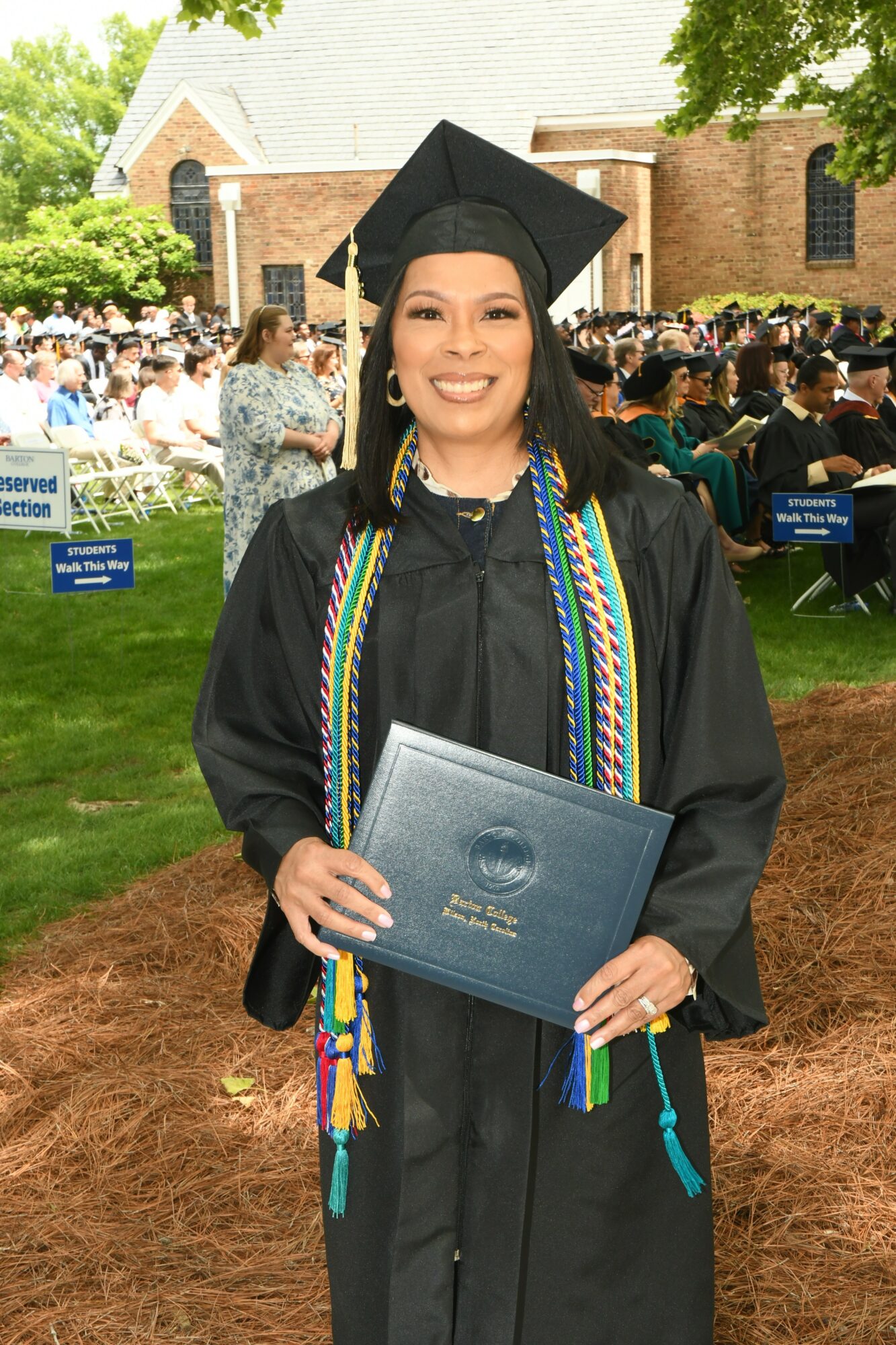 Woman in graduation cap and gown holding diploma outdoors, smiling, with a crowd and a brick building in background.