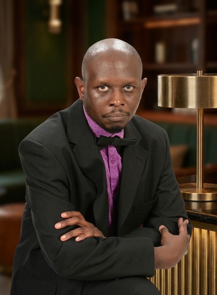 Man with a bald head and serious expression, wearing a black suit, purple shirt, and bow tie, sitting indoors.