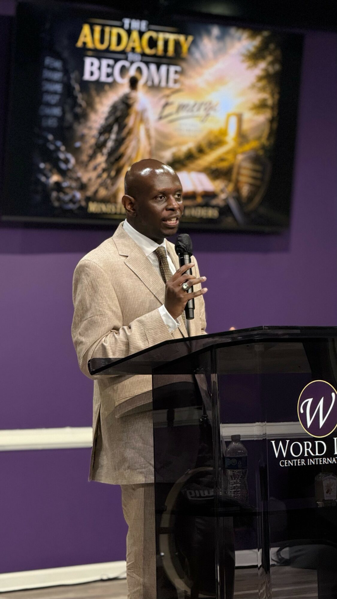 Man in beige suit speaking at a podium with a microphone, in front of a screen displaying a promotional image for 'The Audacity to Become'.