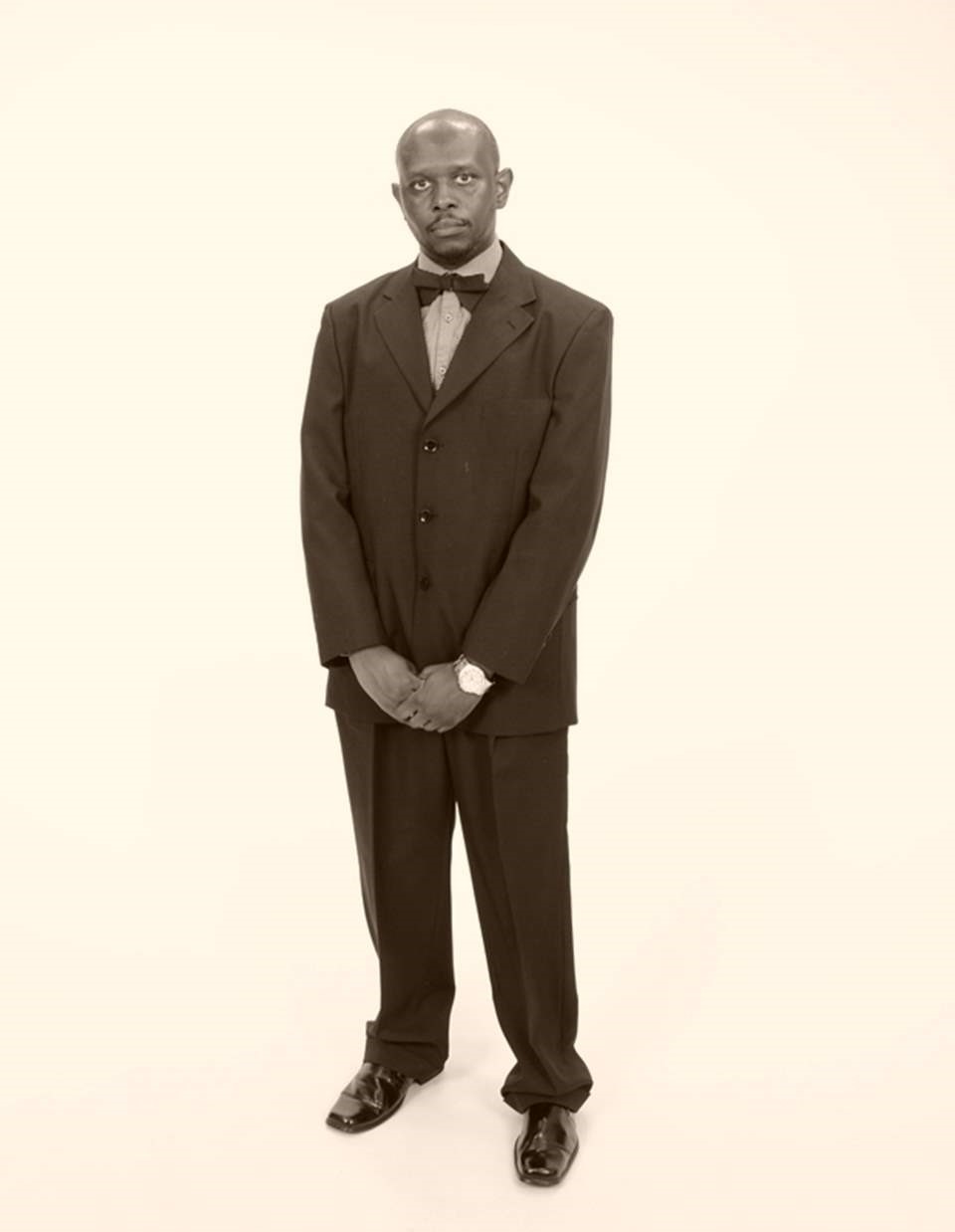 Man in formal suit with bow tie standing with hands clasped, facing forward, against a plain background.