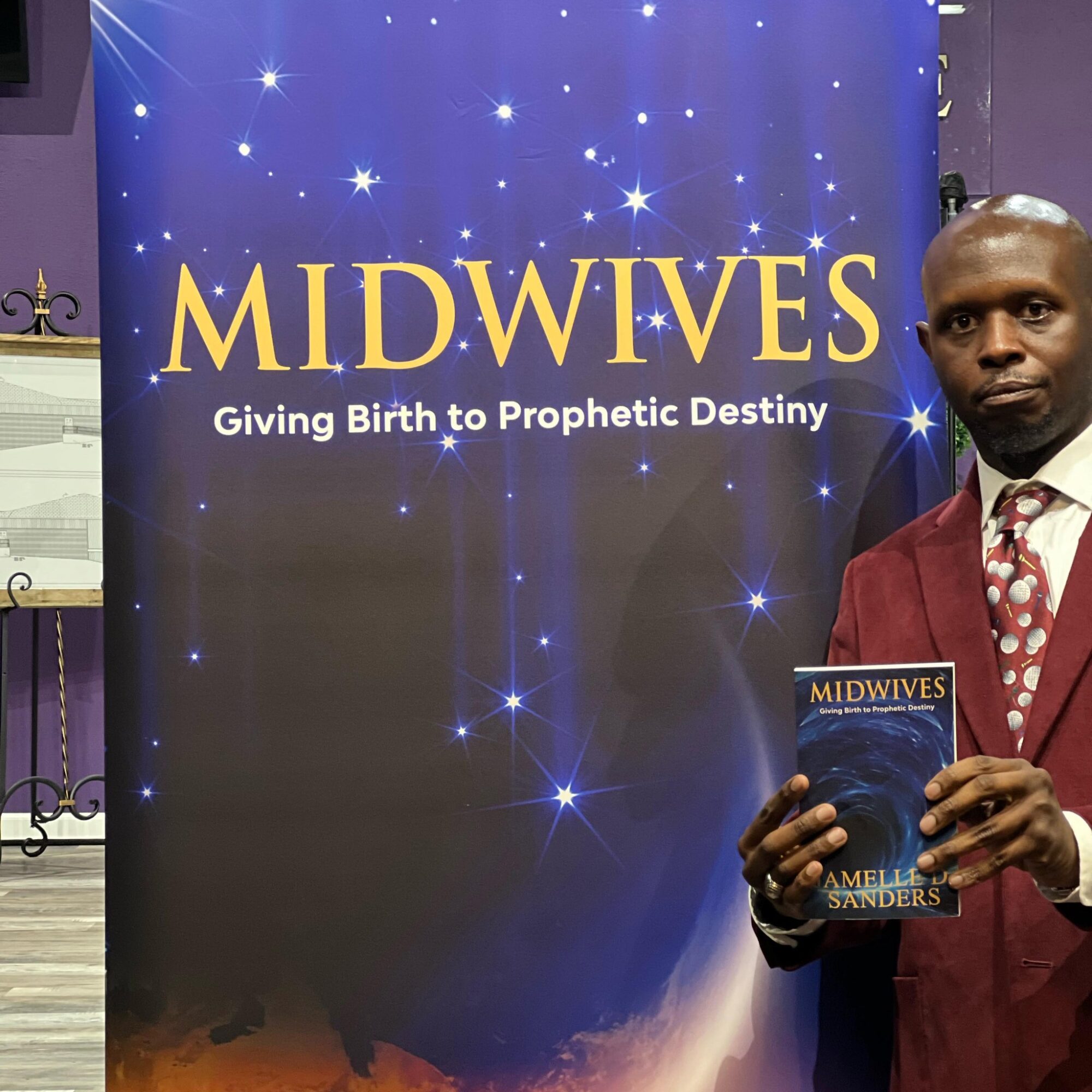 Man in red suit holding a book in front of a blue banner with stars and the text 'MIDWIVES Giving Birth to Prophetic Destiny'.