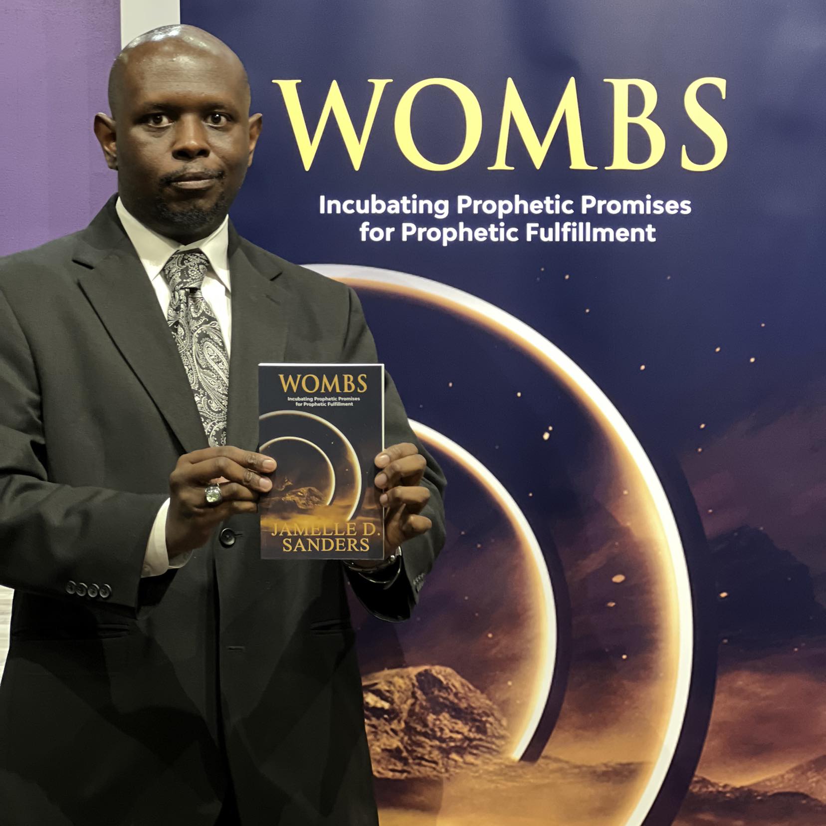 Man in suit holding a book titled 'WOMBS' in front of a space-themed backdrop with planets and stars.