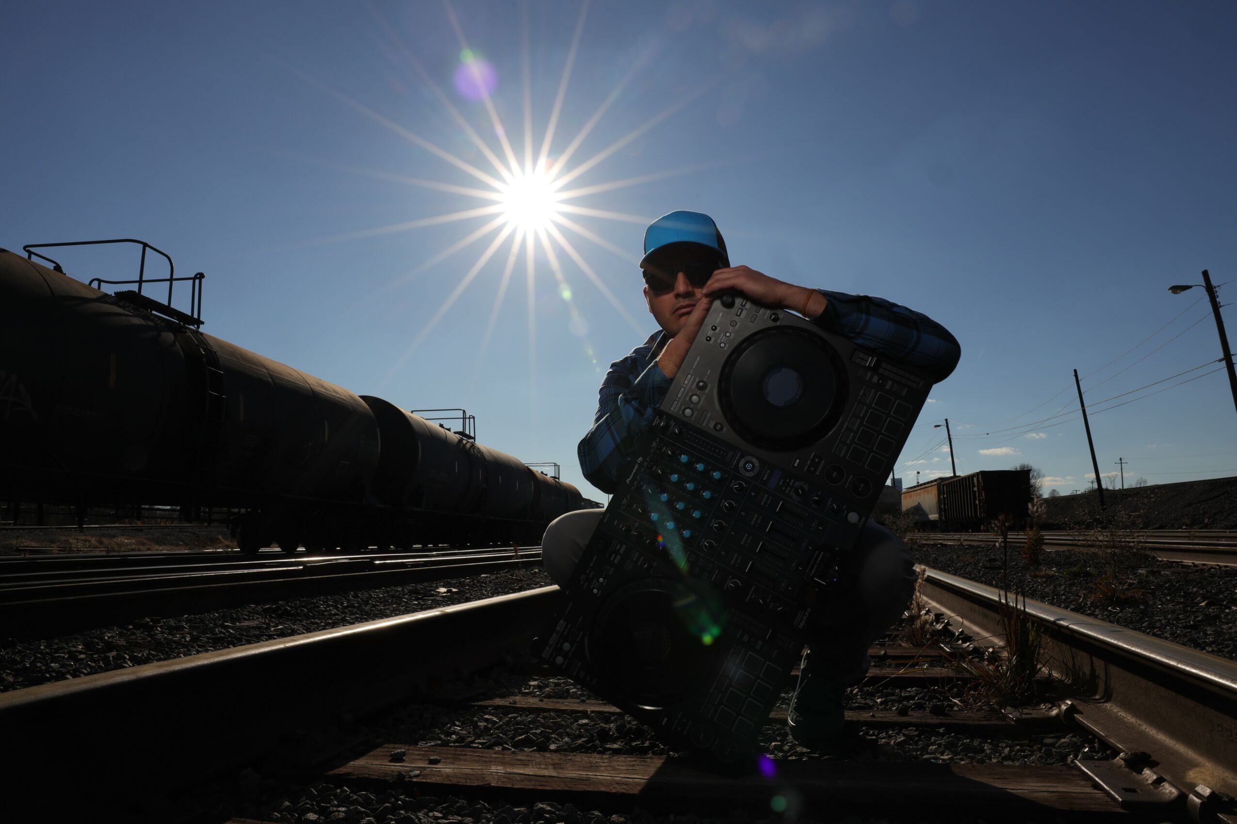 Person crouching on train tracks holding a speaker, with the sun shining brightly in the sky behind them.