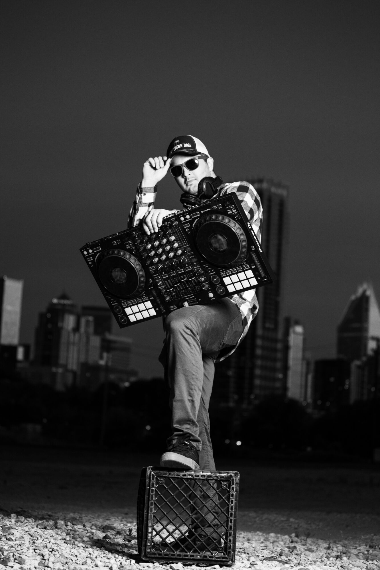 Person wearing sunglasses and headphones, holding DJ equipment, standing on a crate in an urban setting, black and white.