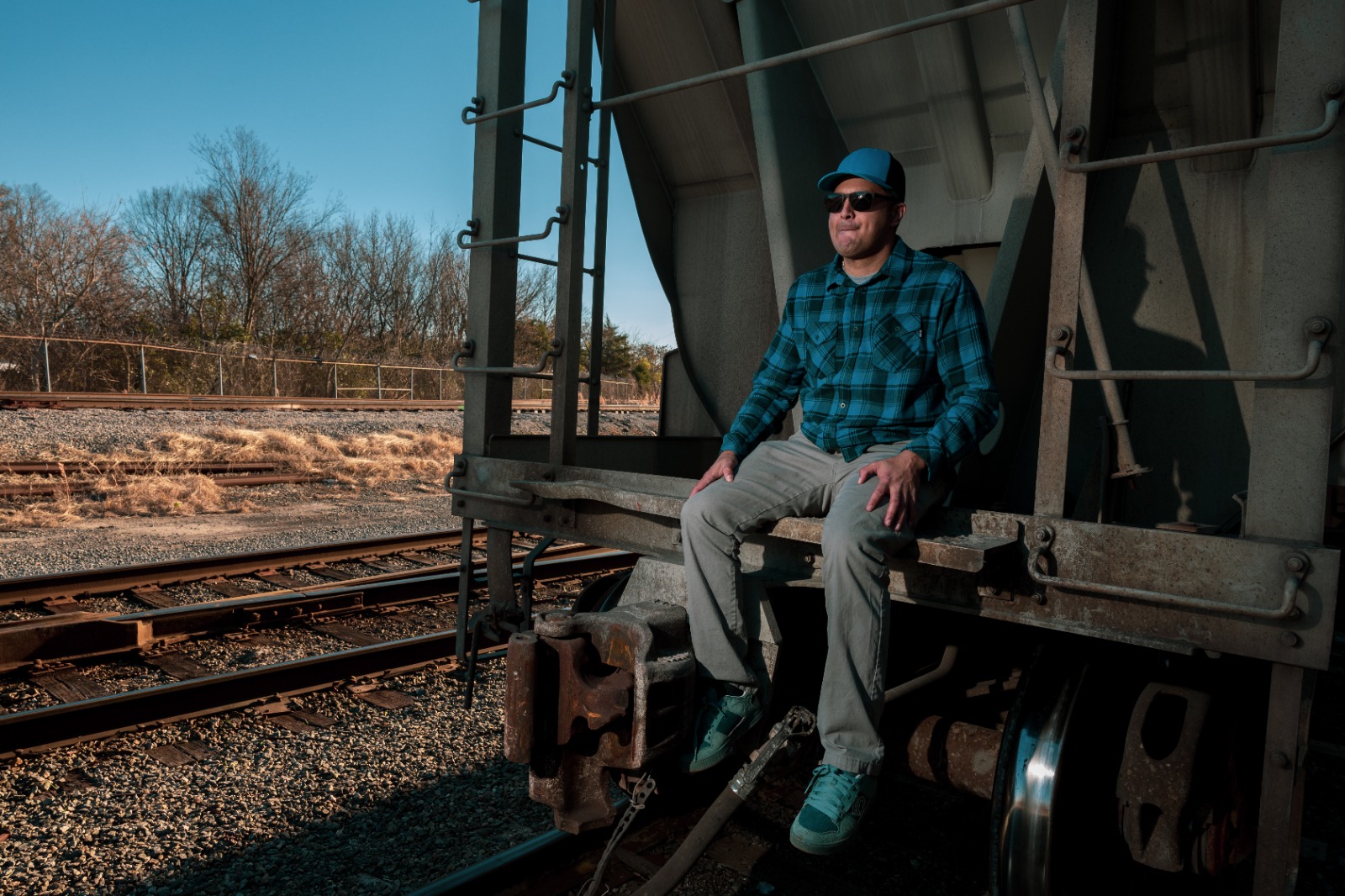 Man sitting on a train car, wearing sunglasses, a blue plaid shirt, and gray pants, outdoors on a sunny day.