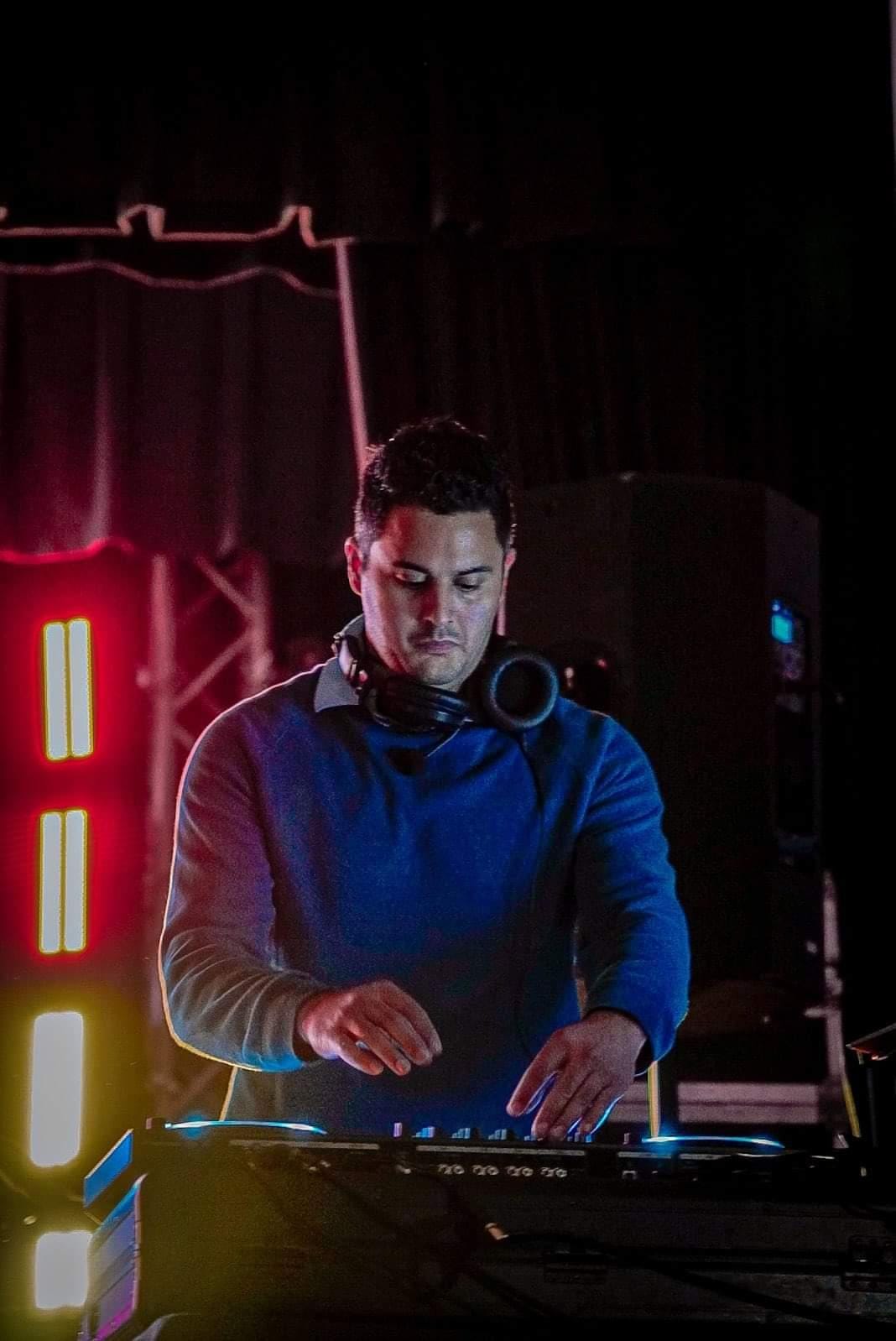 DJ wearing a blue hoodie, focused on mixing music at a nightclub with colorful lights and equipment.