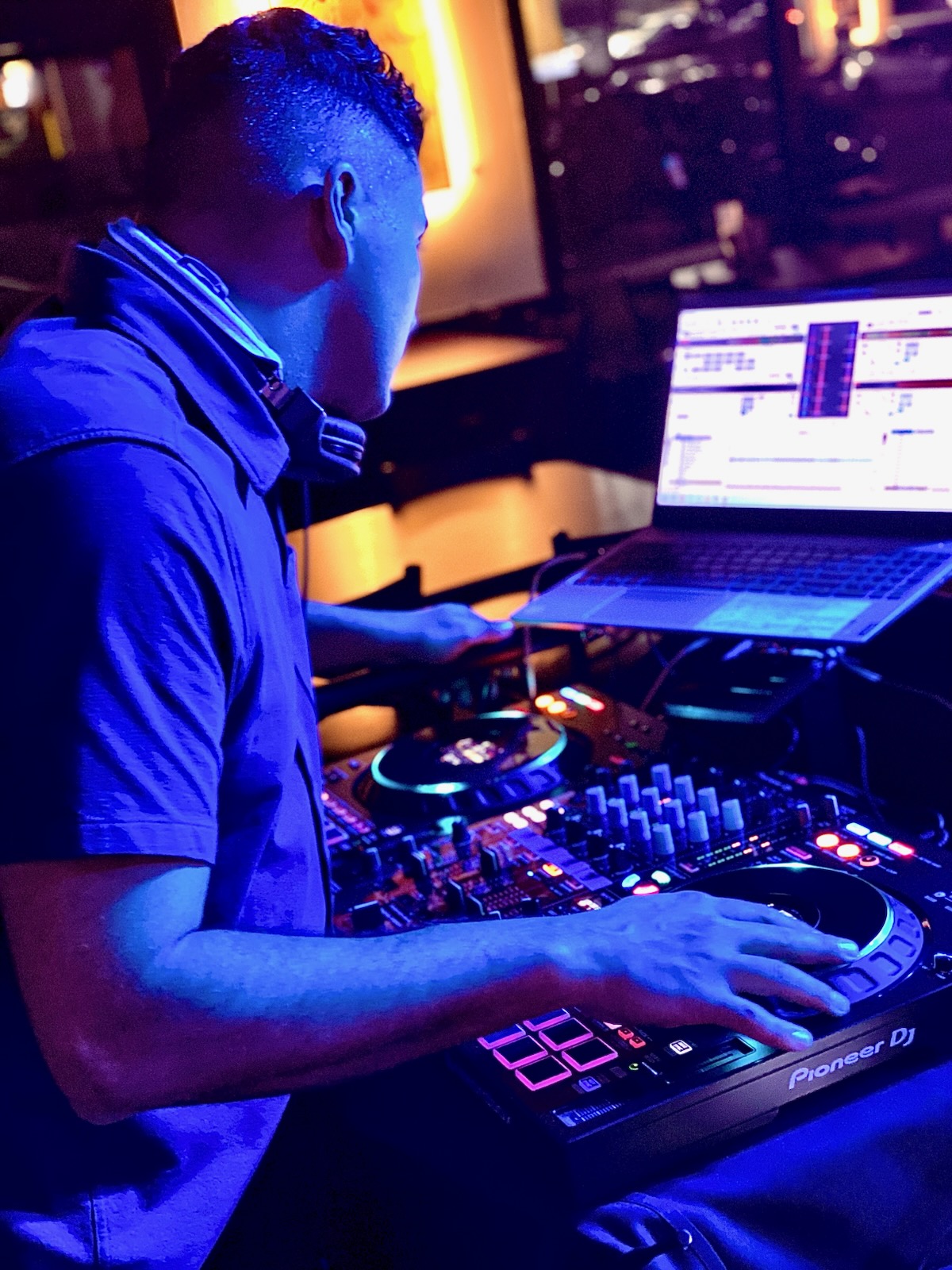 Person DJing with equipment and laptop in a dimly lit environment, illuminated by colorful lights.
