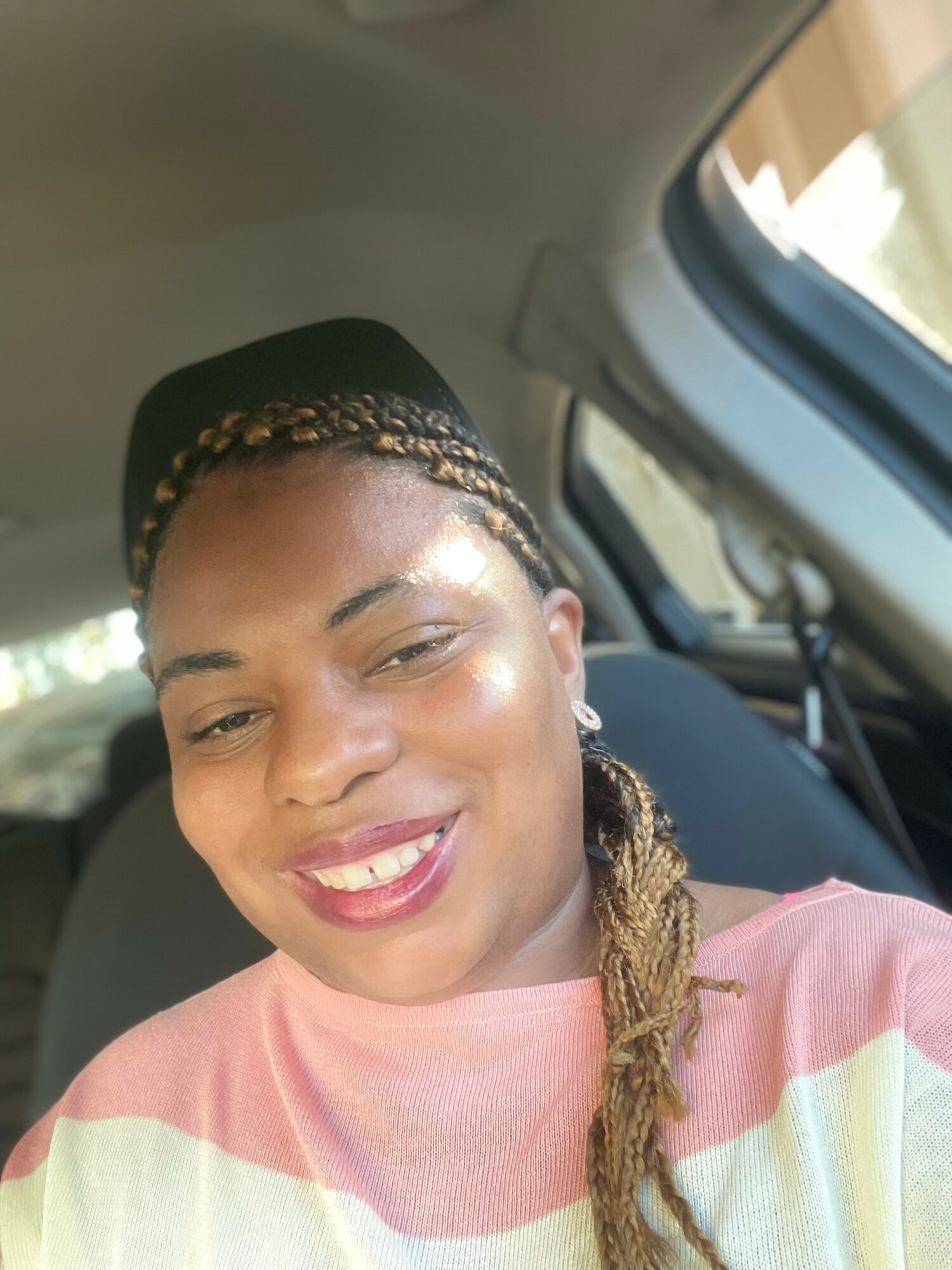Smiling woman with braided hair and a headband, sitting in a car, wearing a pink and white striped top.