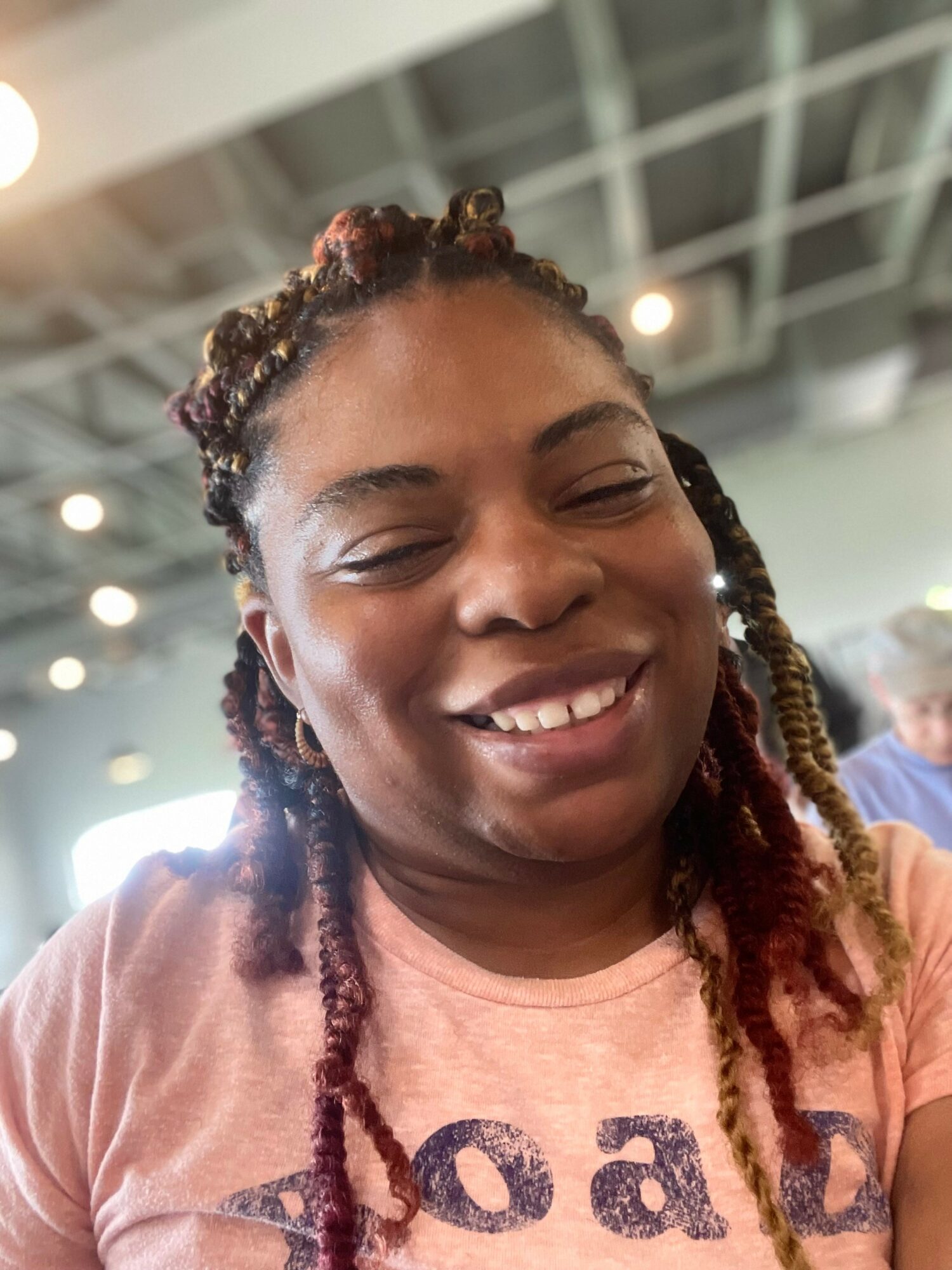 Close-up of smiling woman with braided hair indoors, ceiling with lights visible in background.