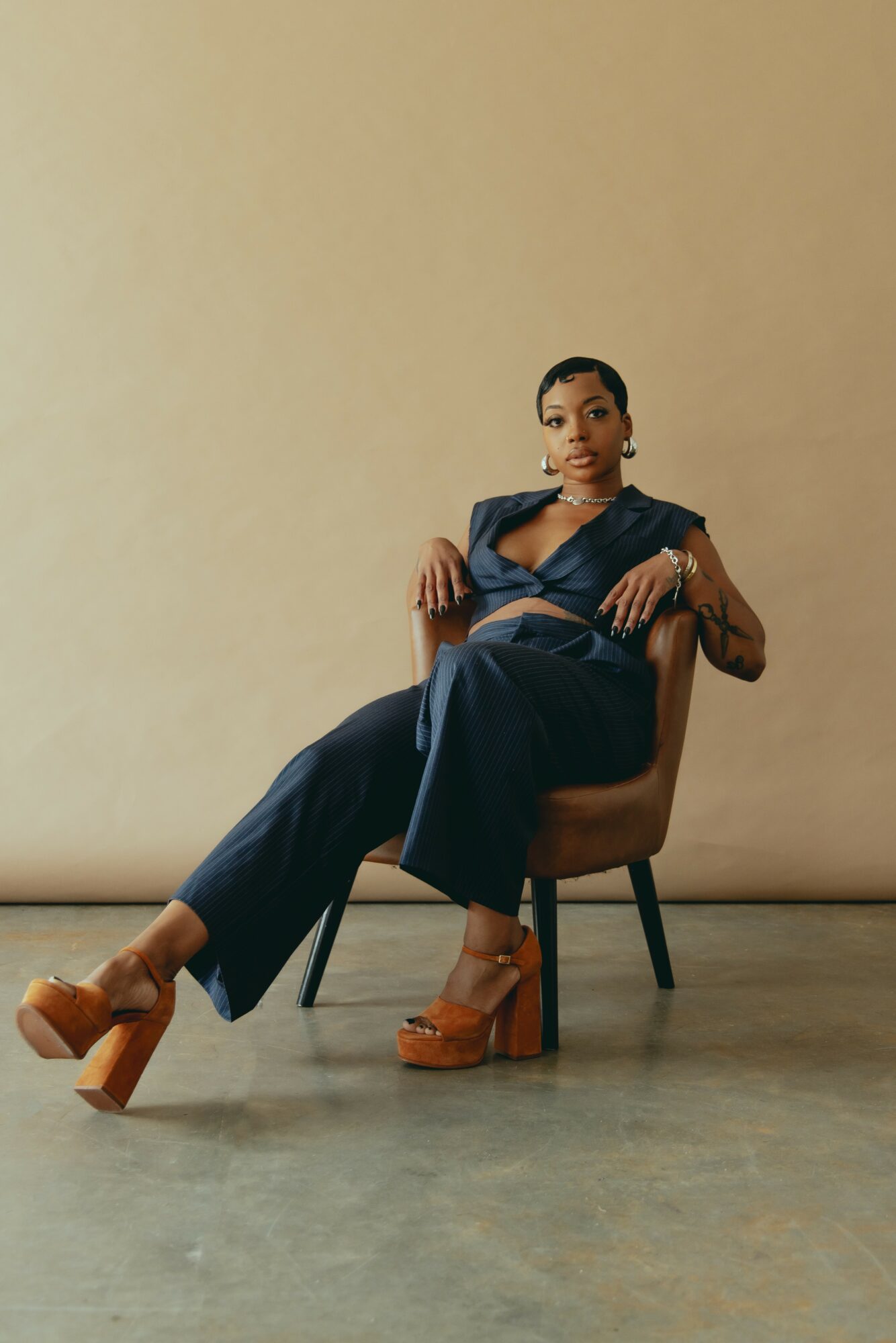 Woman sitting on a chair with legs crossed, wearing a dark outfit and platform shoes, against a plain beige background.