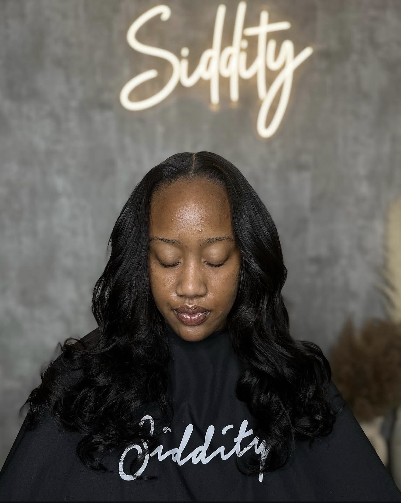 Young woman with long wavy hair and closed eyes, standing in front of a gray background with the word 'Siddity' above her head.