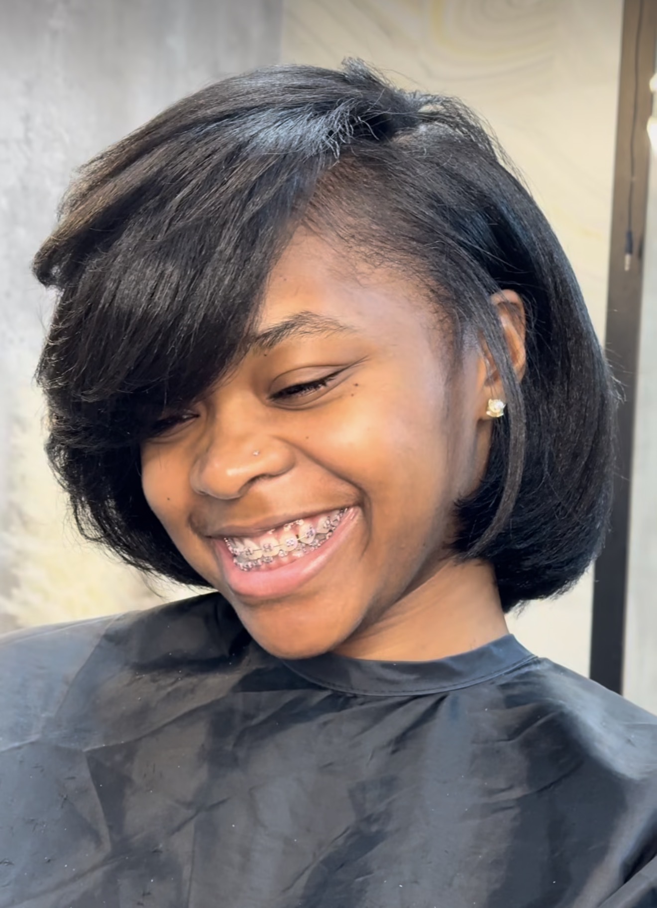 Smiling young girl with braces and a short, layered hairstyle, wearing earrings and a black cape, looking down.