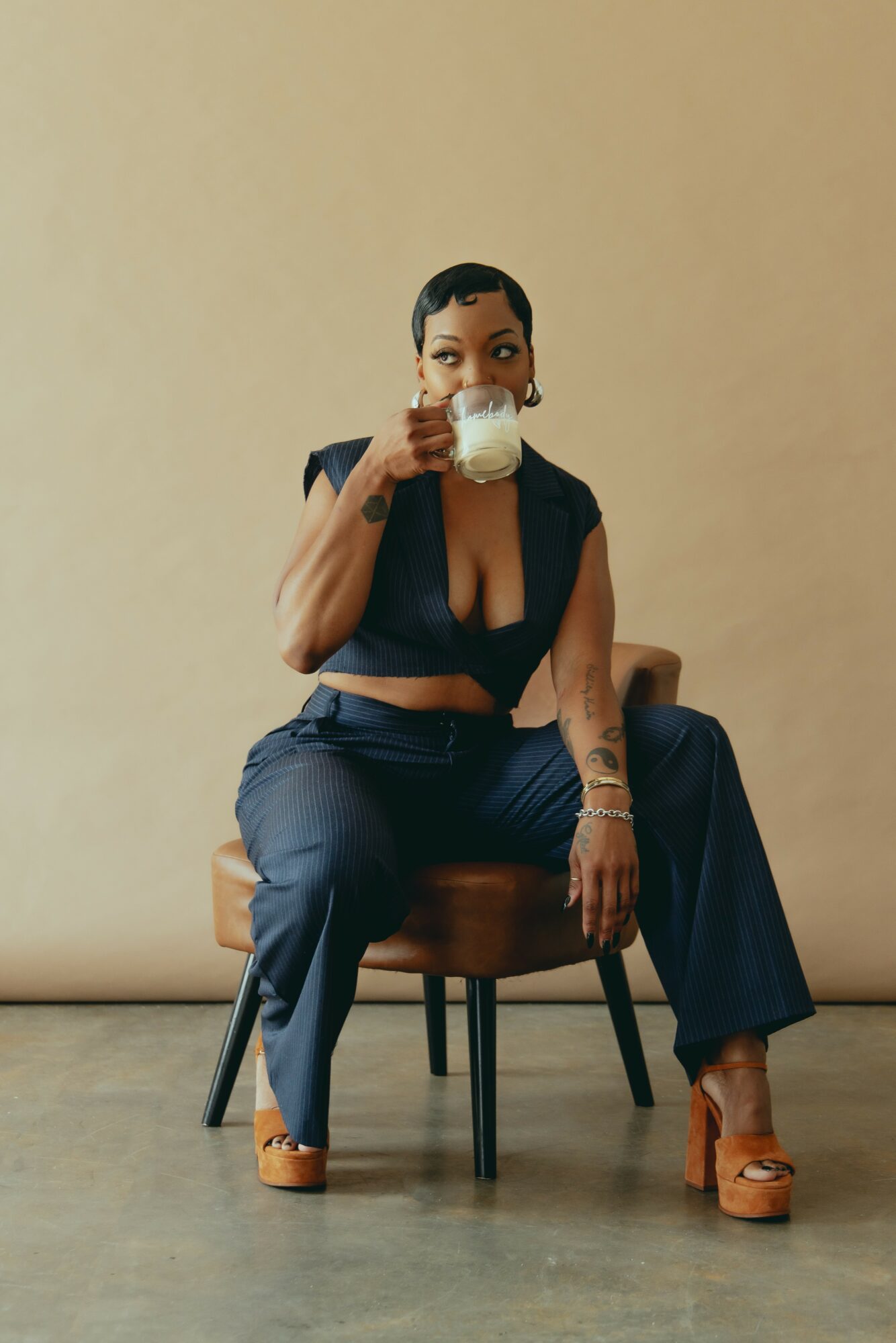 Woman sitting on a chair, drinking from a glass, wearing a black top and dark pants, against a plain beige background.