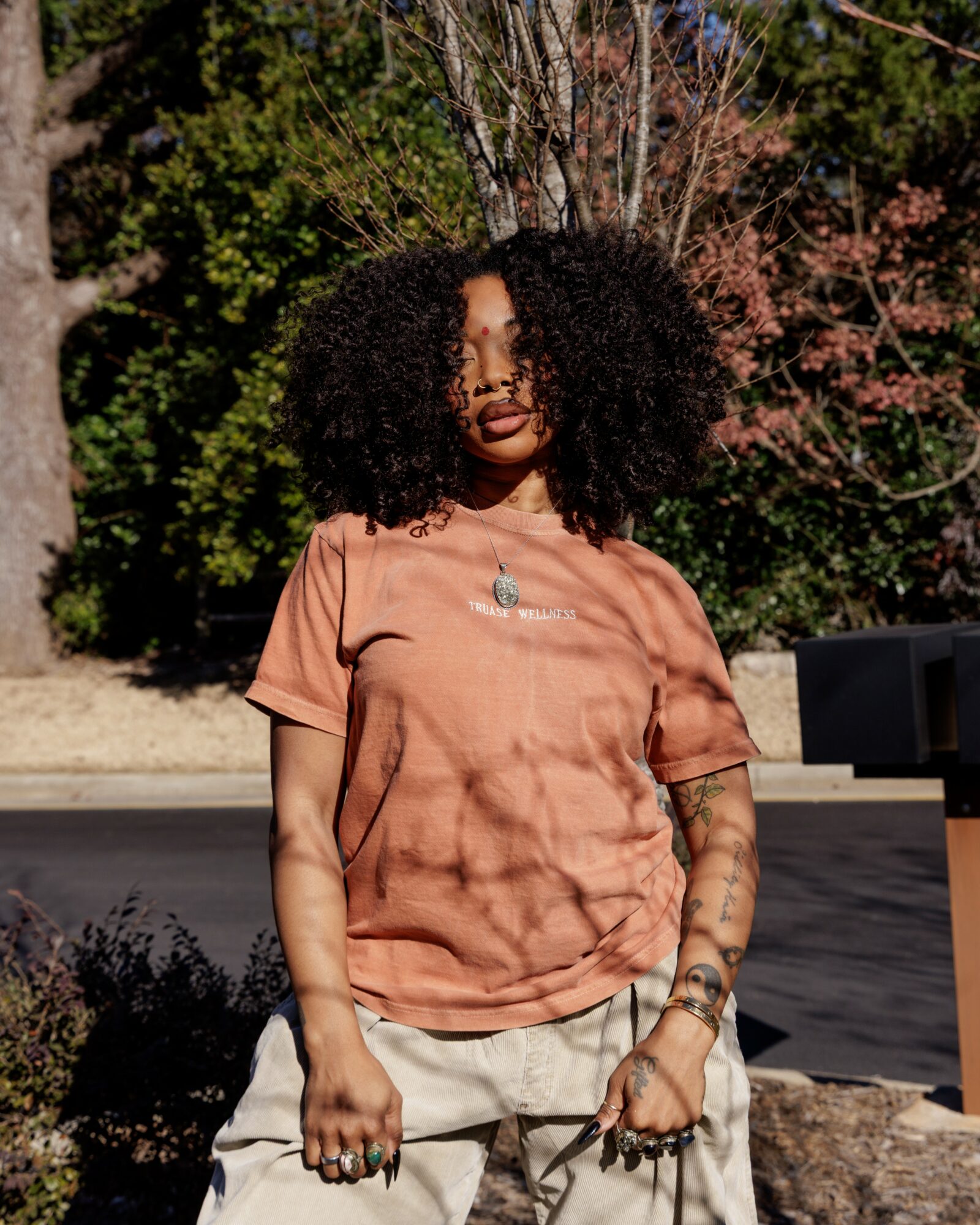 Woman with curly hair wearing a brown T-shirt and light-colored pants outdoors, trees in background.