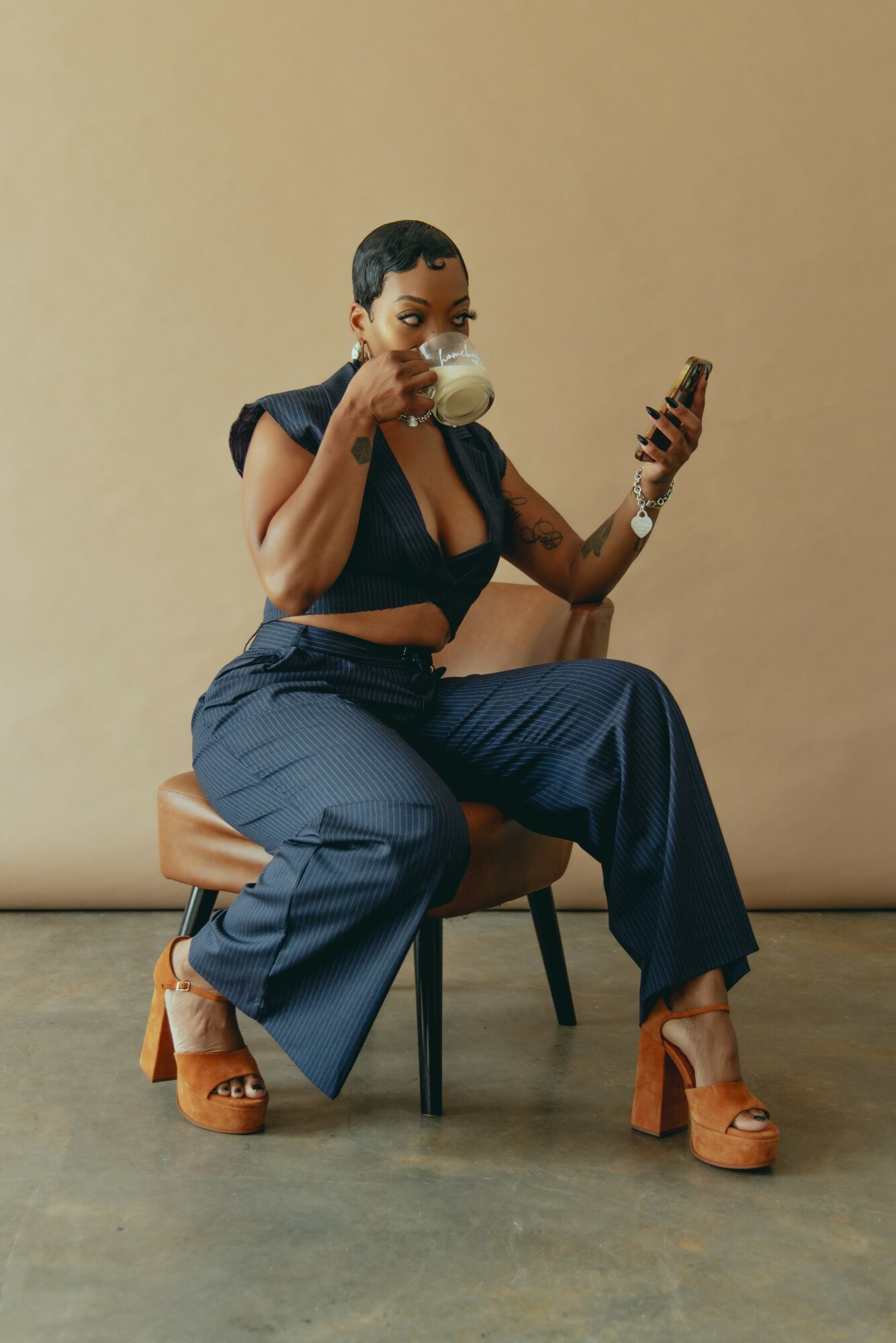 Woman sitting on a chair, holding a cup and a phone, wearing a dark top, wide pants, and platform shoes.